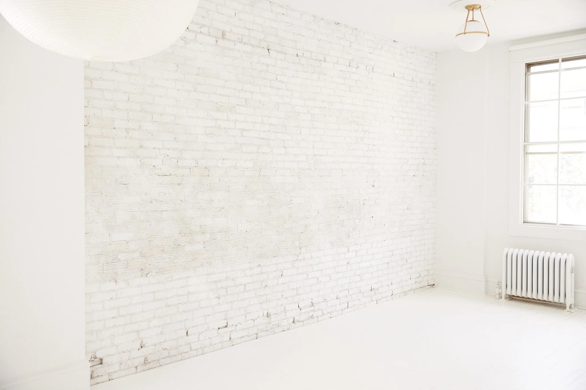 Empty room with white brick wall, white floor, window, and radiator, bright natural light, minimalist aesthetic.