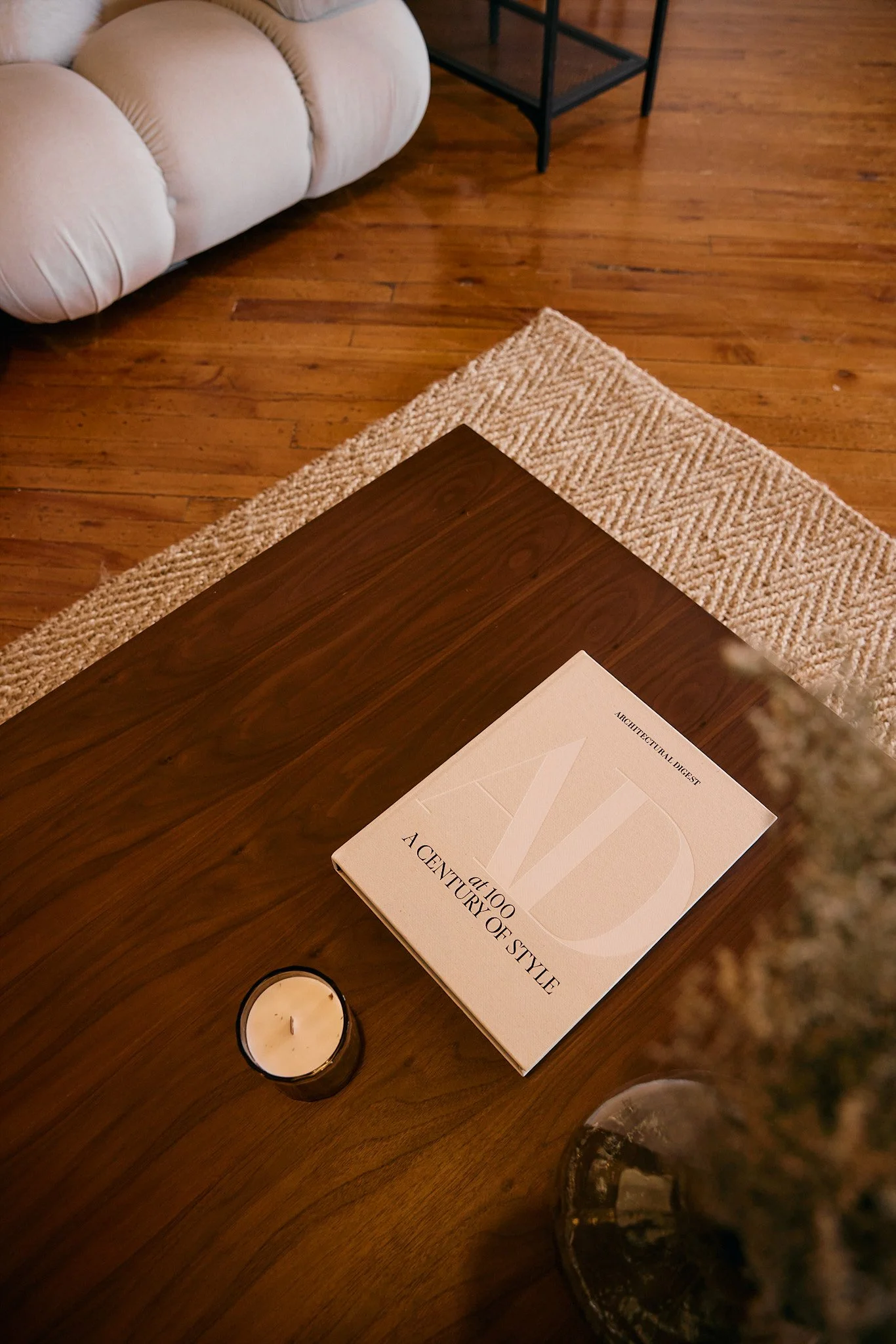 Close-up of a wooden table with a book titled 'A Century of Style' on it, a lit candle, a candle holder, a rug, part of a sofa, and a small table in the background.