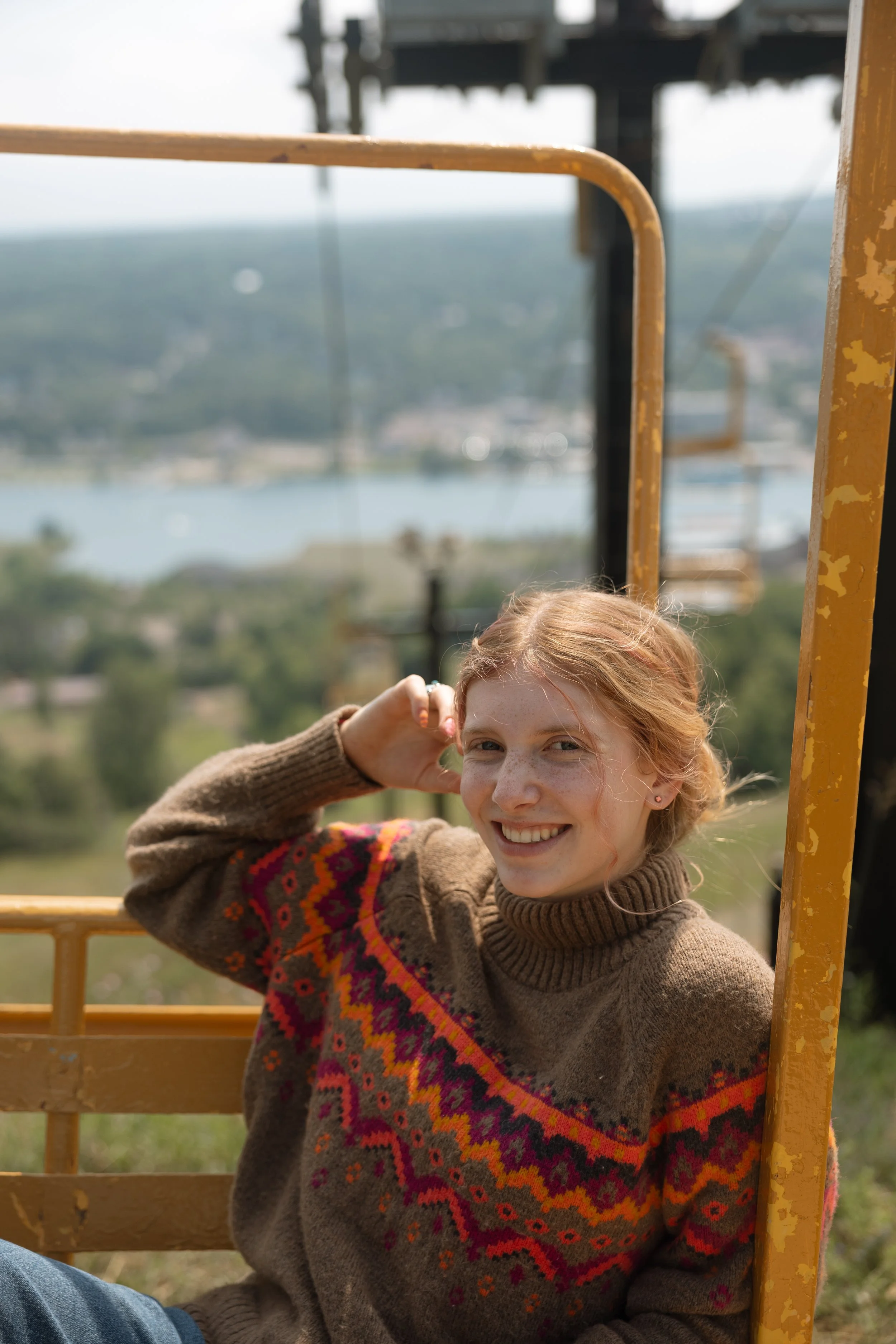 A young woman with red hair and freckles smiling while sitting on a yellow metal structure outdoors, with a distant view of a river and trees in the background. Mt. Ripley ski lift 