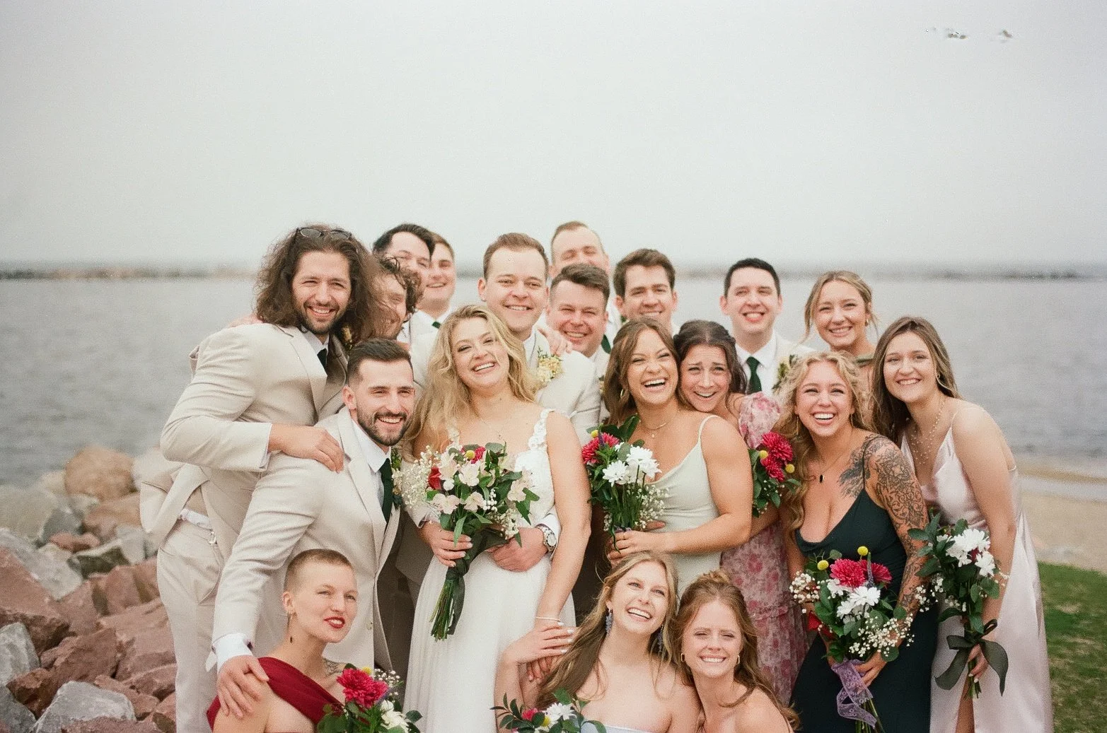 A large group of people dressed in formal wedding attire, standing outdoors near a body of water, smiling and holding bouquets, celebrating a wedding.