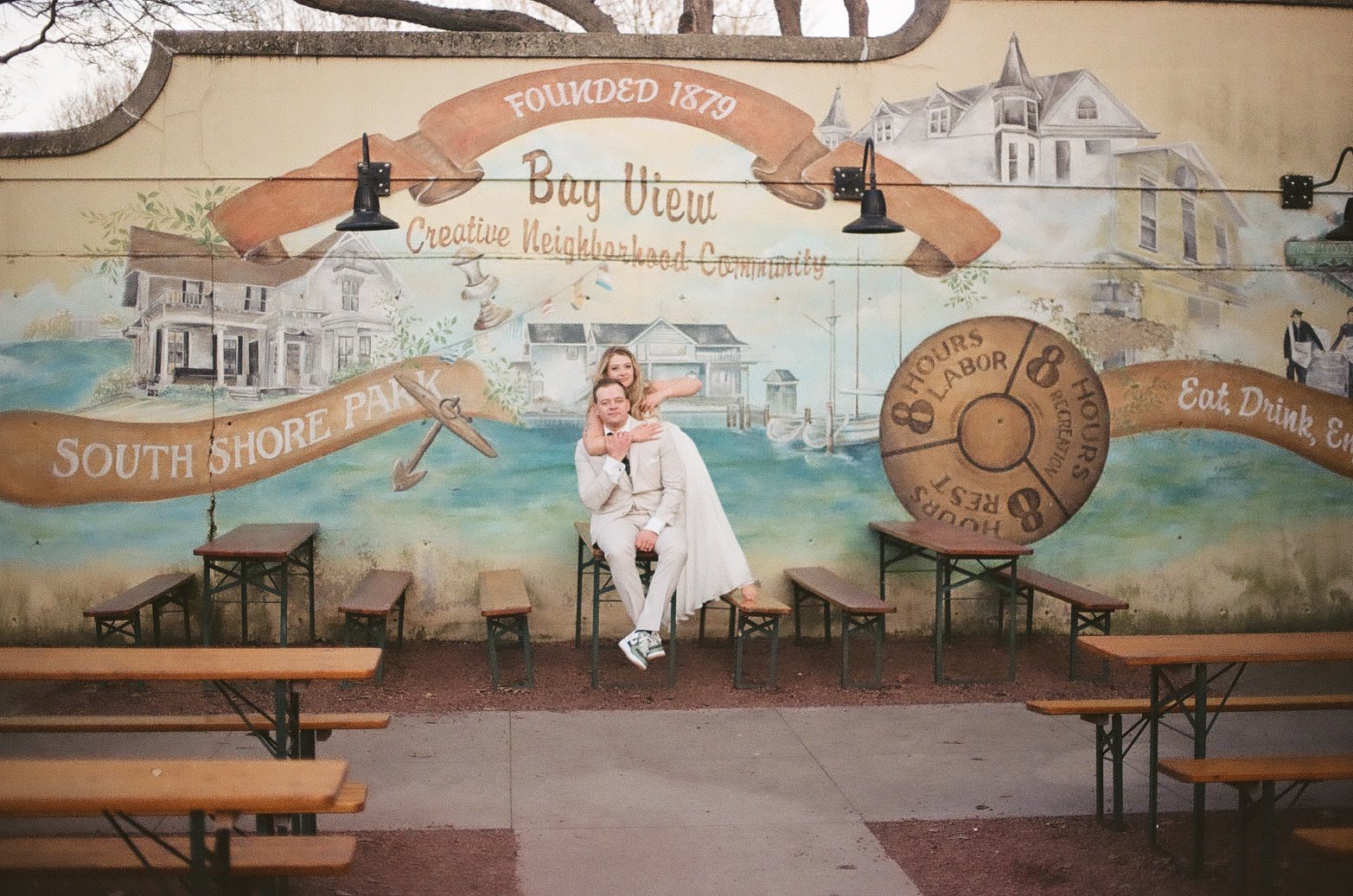 A mural on a wall advertising Bay View, a neighborhood founded in 1870, featuring houses, boats, and an anchor with the text 'South Shore Park.' In front, a young couple dressed in white is sitting on a bench, with the woman hugging the man, against the mural backdrop.