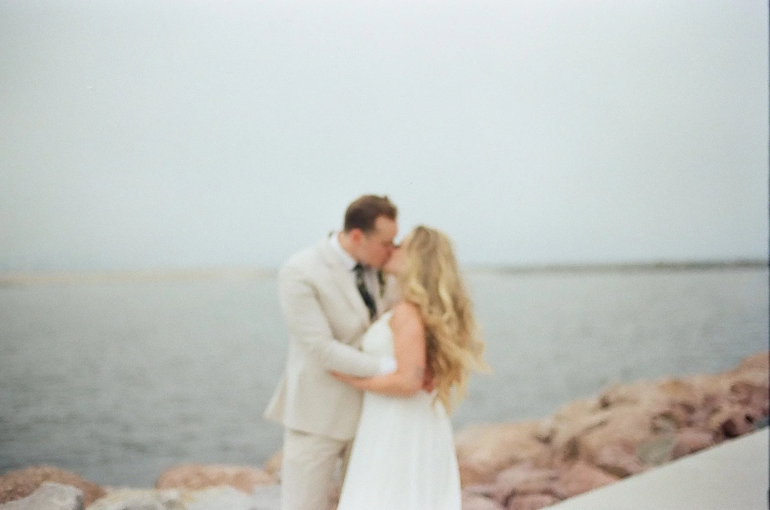 A couple in wedding attire sharing a kiss by the water on a rocky Milwaukee shoreline.
