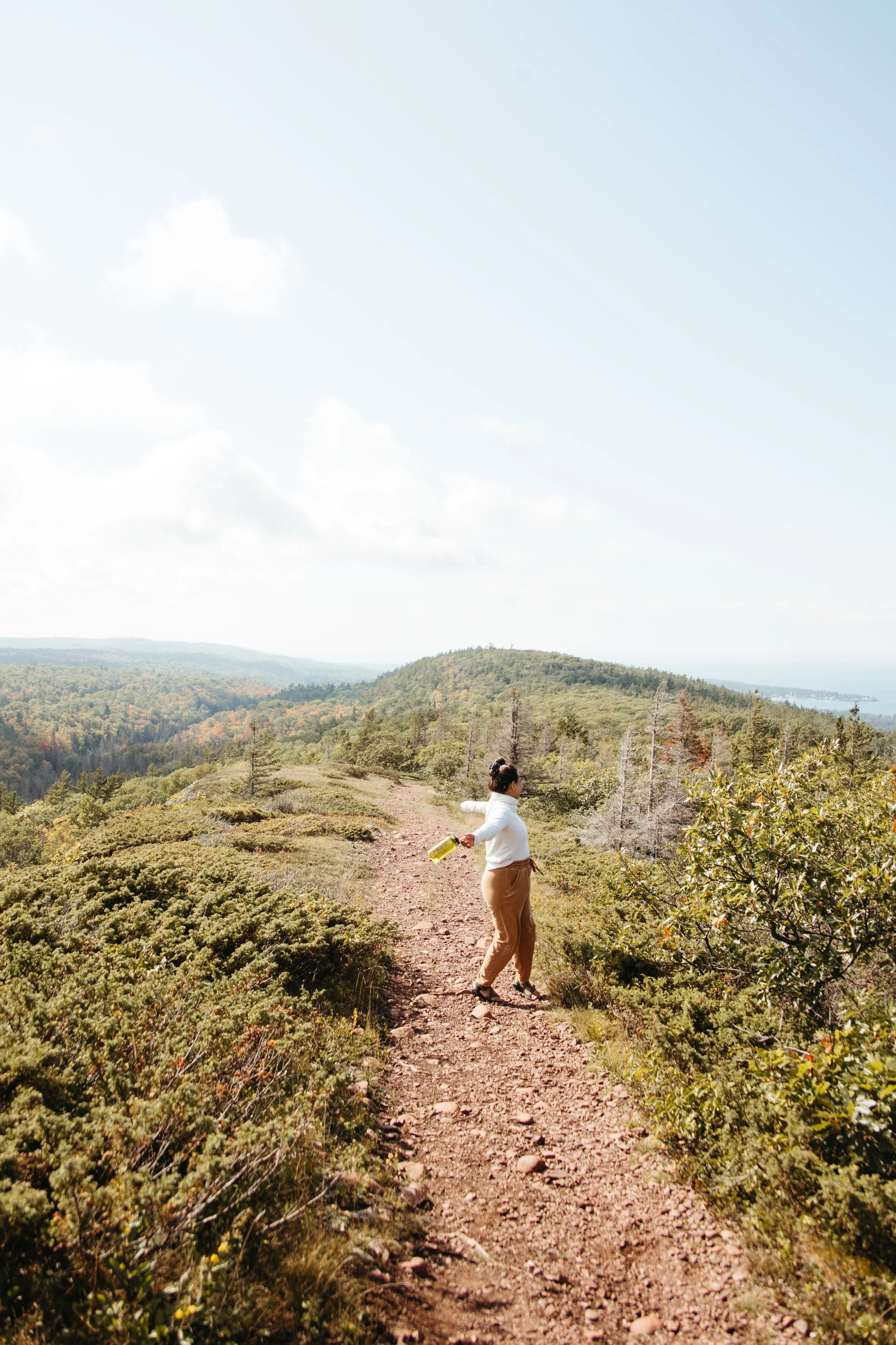 A woman in beige pants and a white jacket walking along a dirt trail in a hilly, forested area with distant hills and partly cloudy sky. Visit Keweenaw