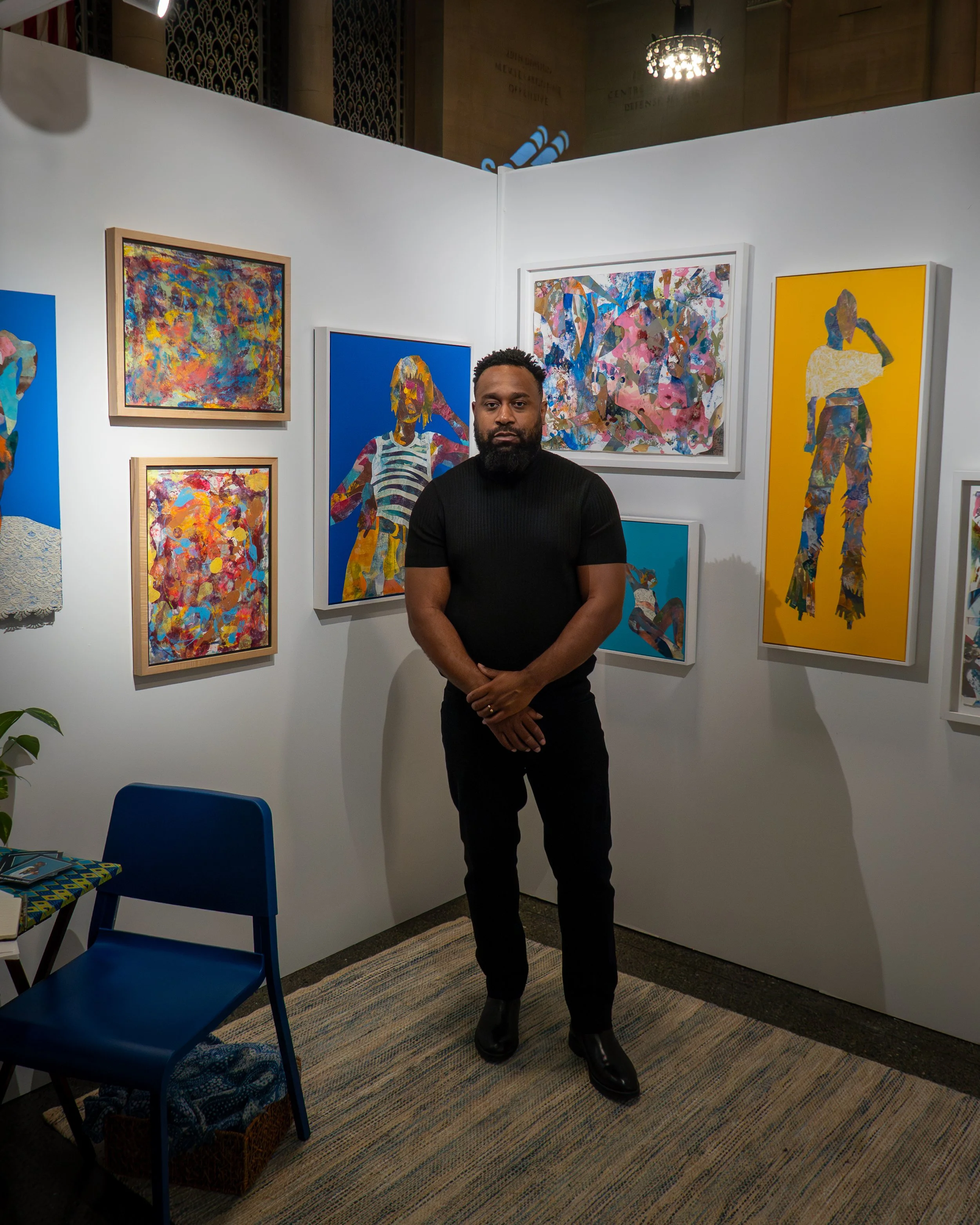 A man with a beard and short hair wearing a black turtleneck and black pants standing in an art gallery with colorful abstract paintings on white walls. There is a blue chair and a woven basket on the floor, and a decorative rug under the man's feet.