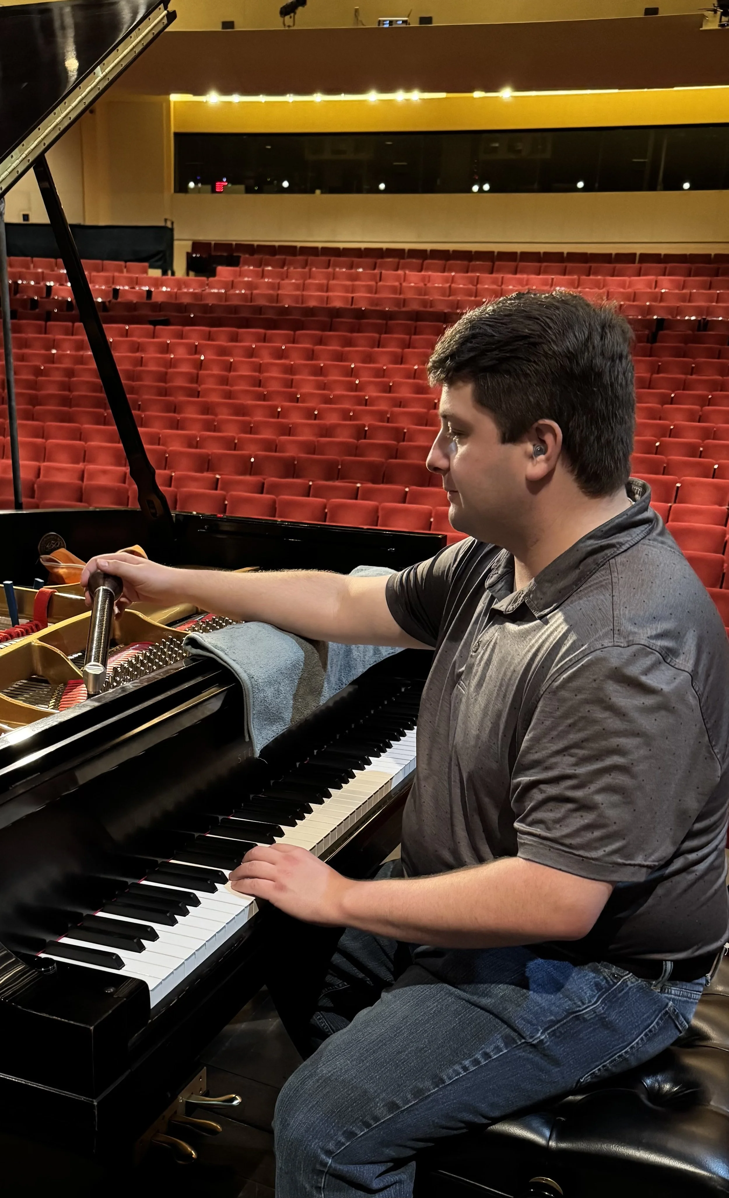 A man sitting at a grand piano on a stage with red seats in the background.