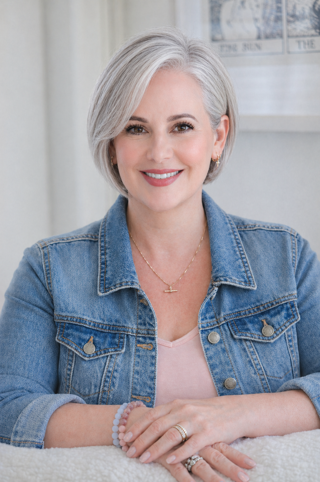 Smiling middle-aged woman with short gray hair, wearing a denim jacket and jewelry, sitting with her hands folded on a table.