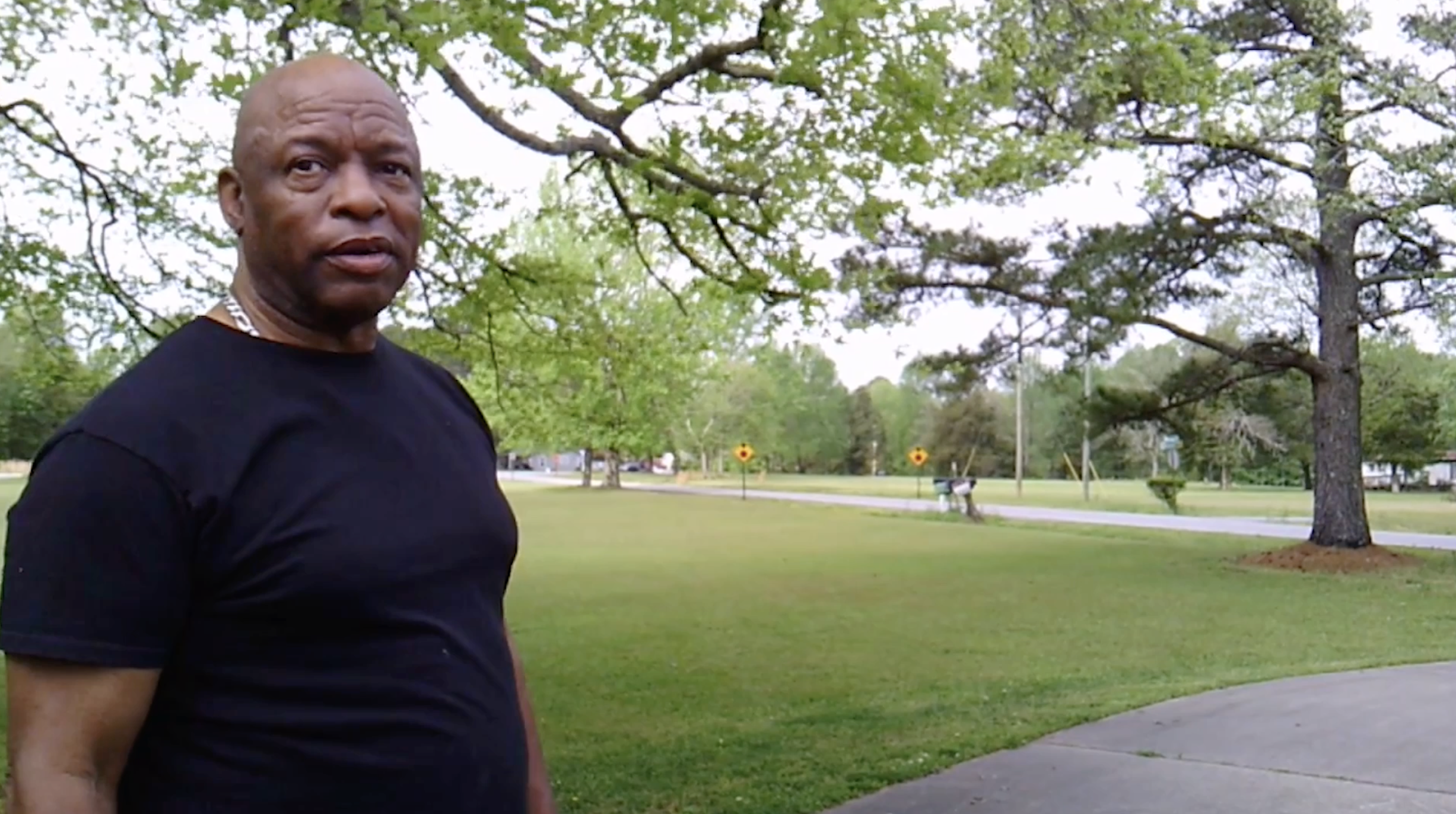A man in a black shirt standing on a sidewalk in a park with trees and grass, and two yellow road signs in the background.