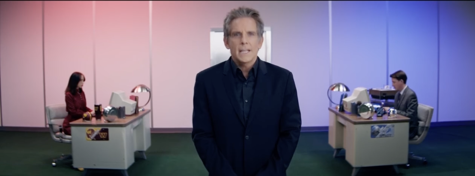 A man in a dark suit stands in front of a colorful office space with pink and blue walls, with a man and woman working at desks in the background.