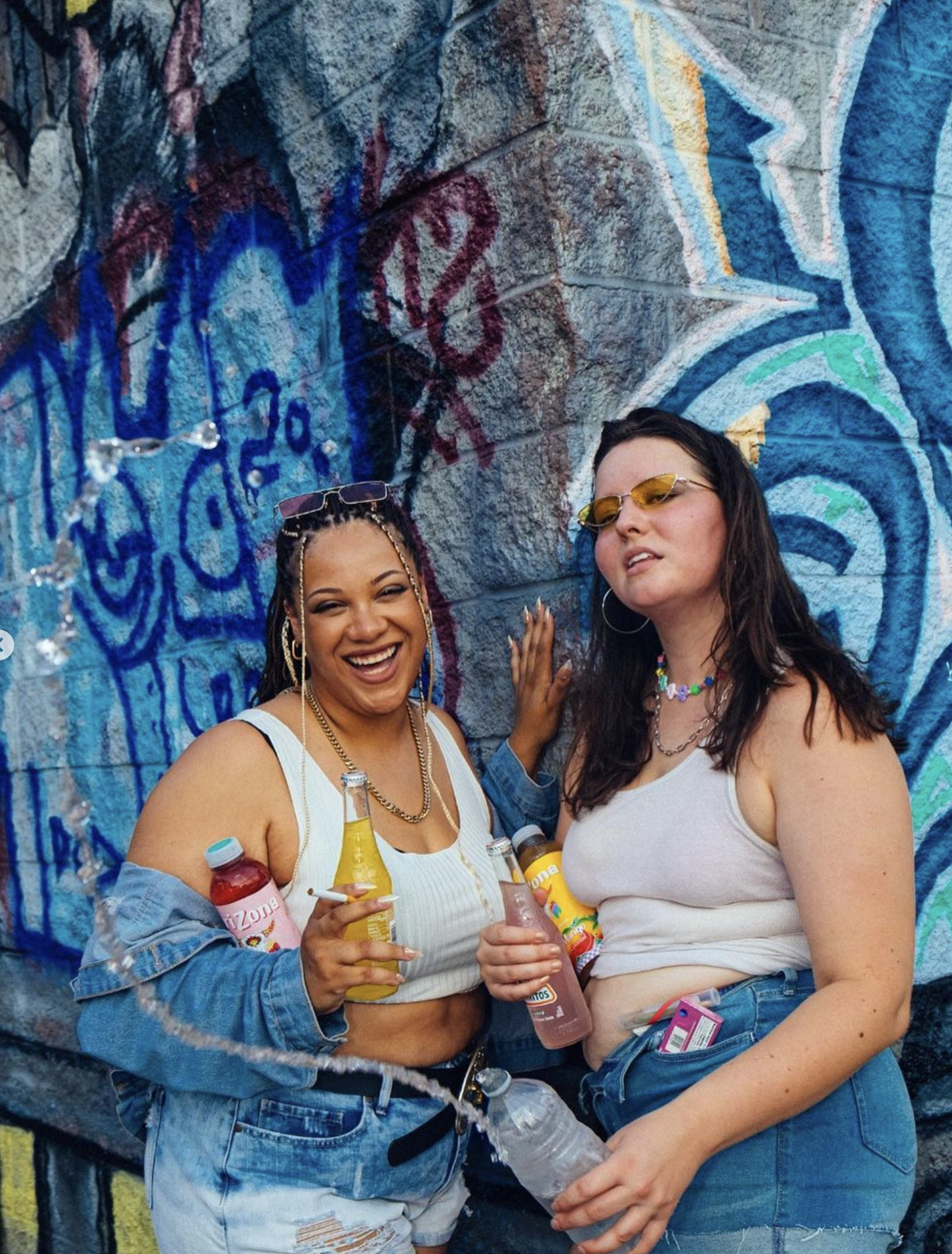 Two young women standing in front of graffiti art on a brick wall, holding drinks and smiling. One has facial jewelry and braided hair, the other wears sunglasses and hoop earrings.