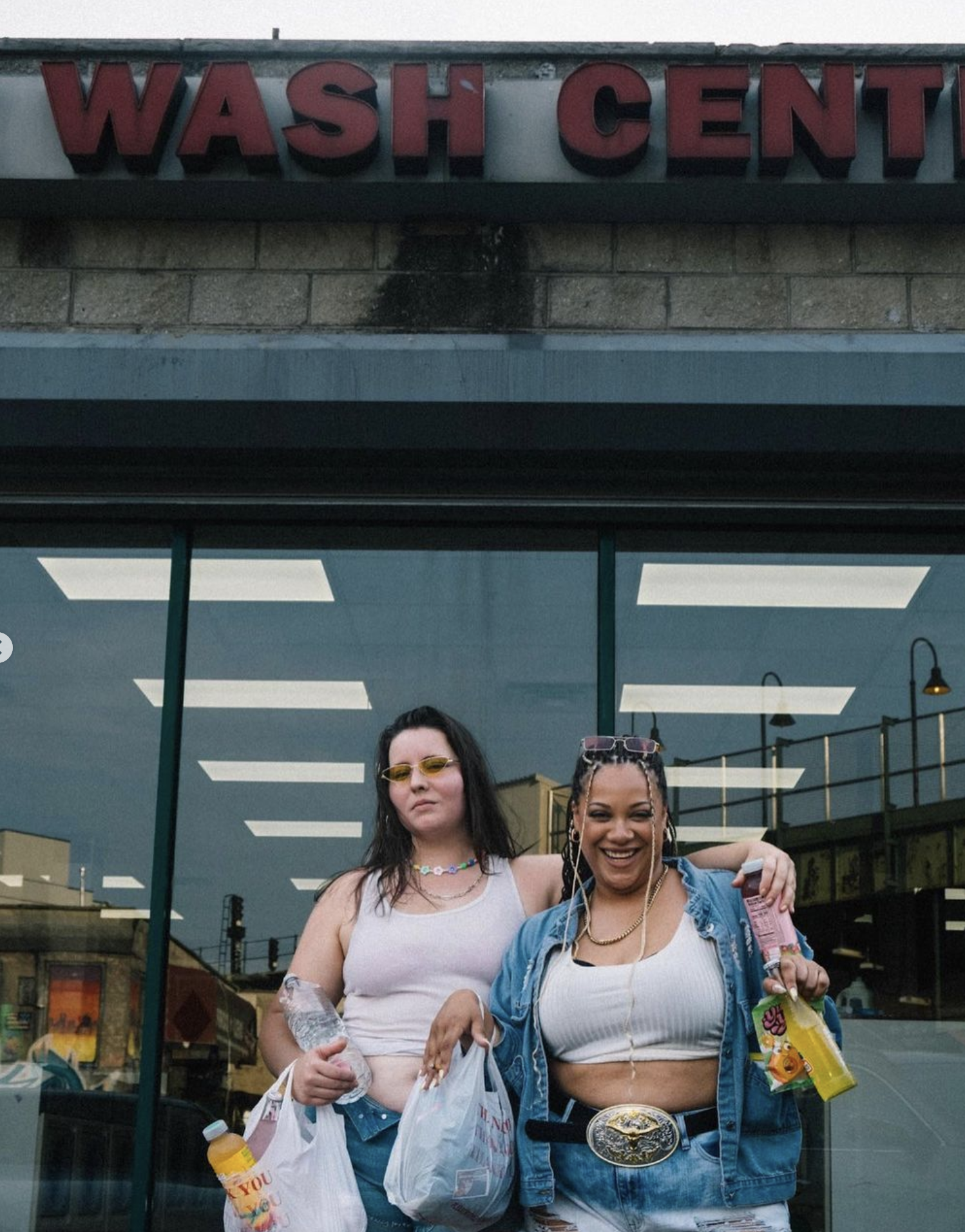 Two women stand outside a laundromat with a sign that reads 'WASH CENT', holding shopping bags and drinks, smiling and enjoying themselves.