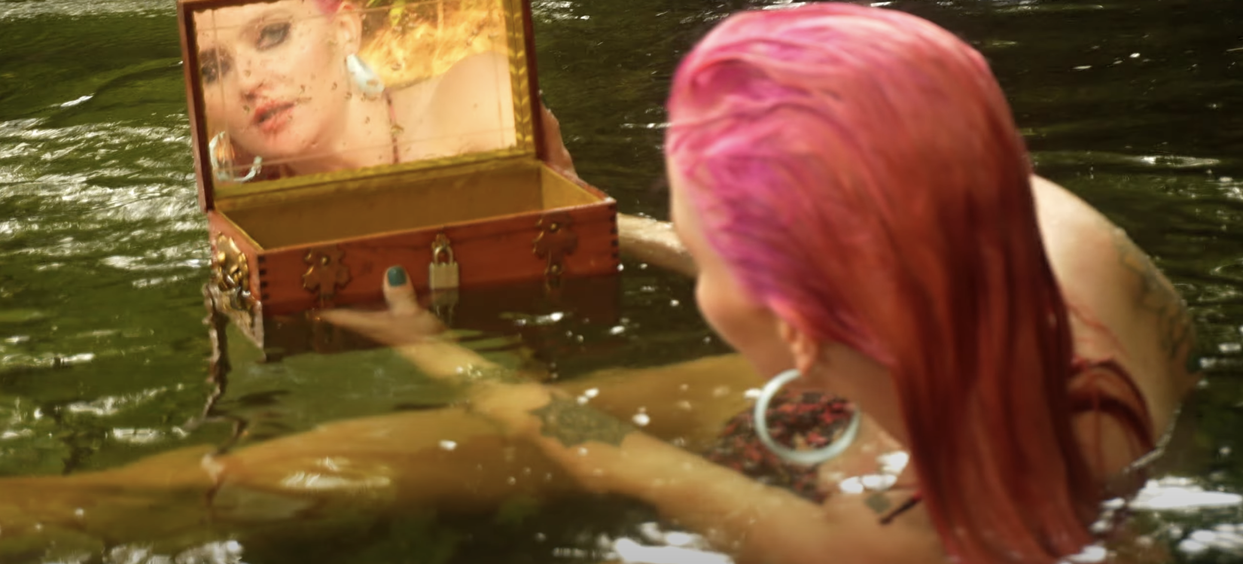 Woman with pink hair looking into a mirror inside a small wooden box in a body of water.