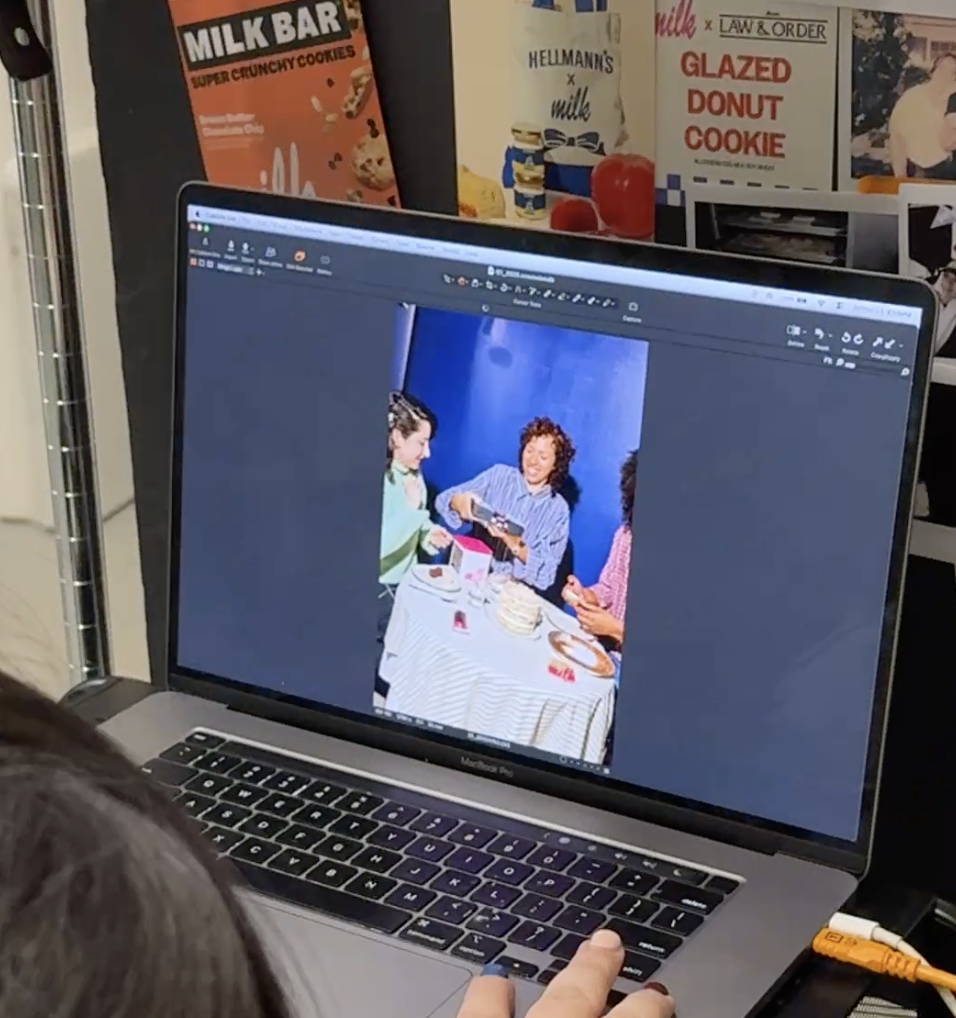 A laptop displaying a photo of three women sitting at a table with birthday cake and snacks, with posters related to baked goods and cookies in the background.
