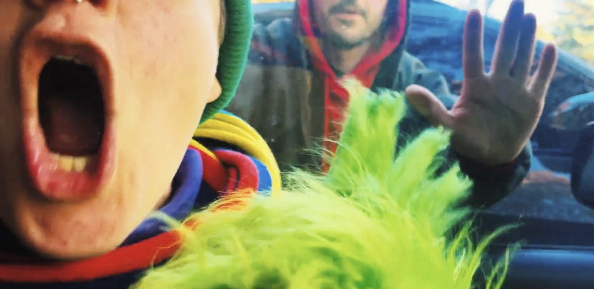 Children in colorful costumes desperately trying to catch a fuzzy green and yellow plush toy in front of a car window