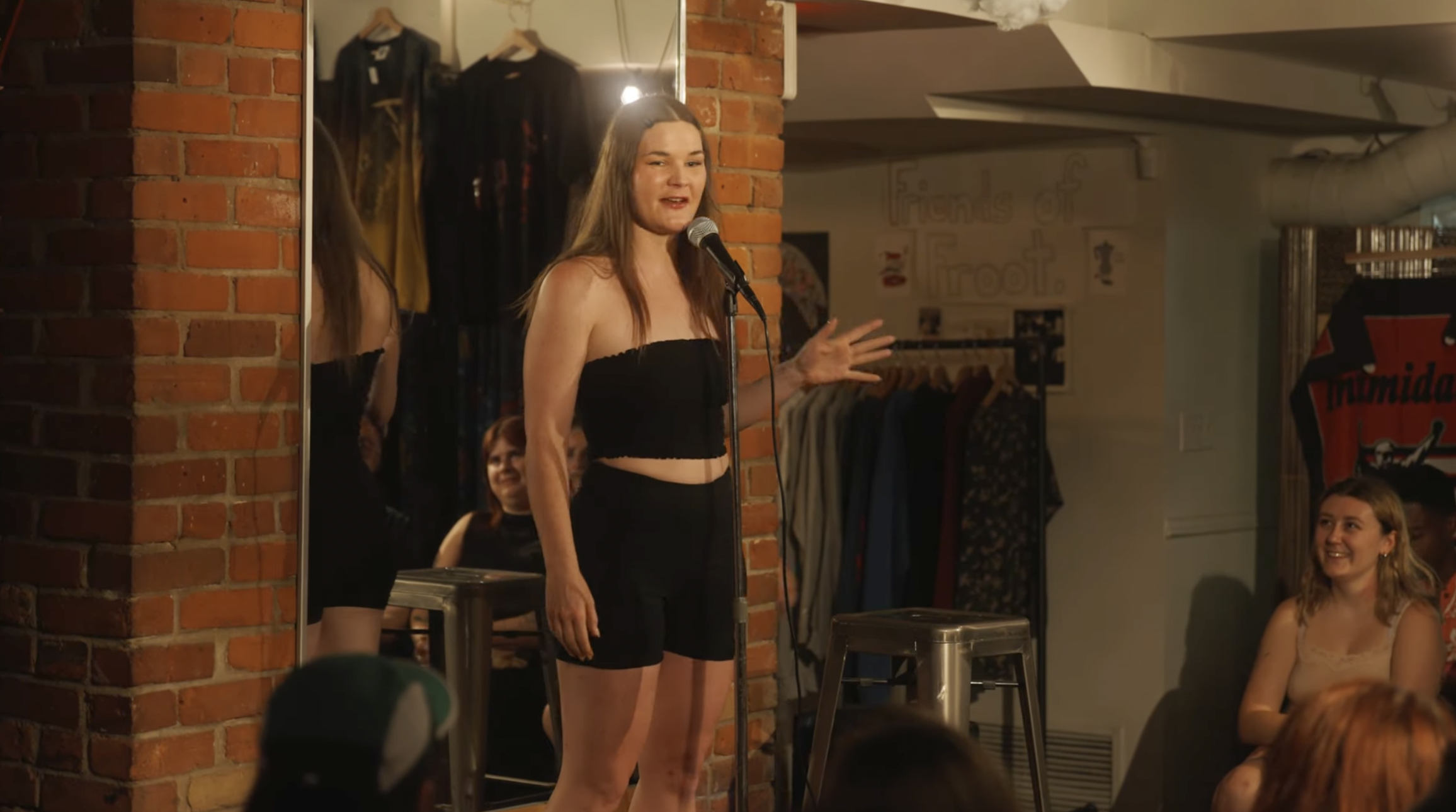 A woman is performing stand-up comedy on stage, standing in front of a microphone, wearing a black strapless top and shorts, with an audience watching and smiling.