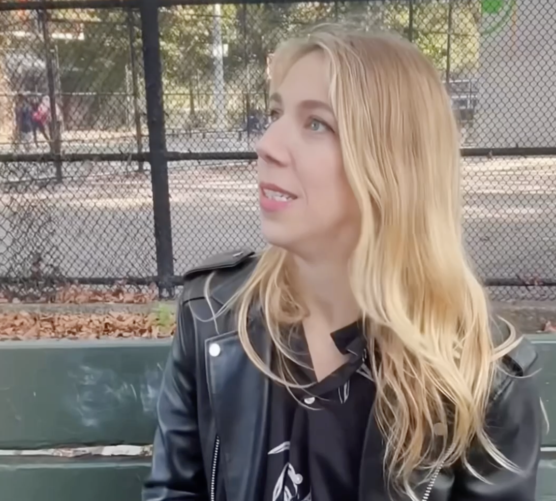 A woman with long blonde hair sitting on a park bench, wearing a black leather jacket, with a chain-link fence and people in the background.
