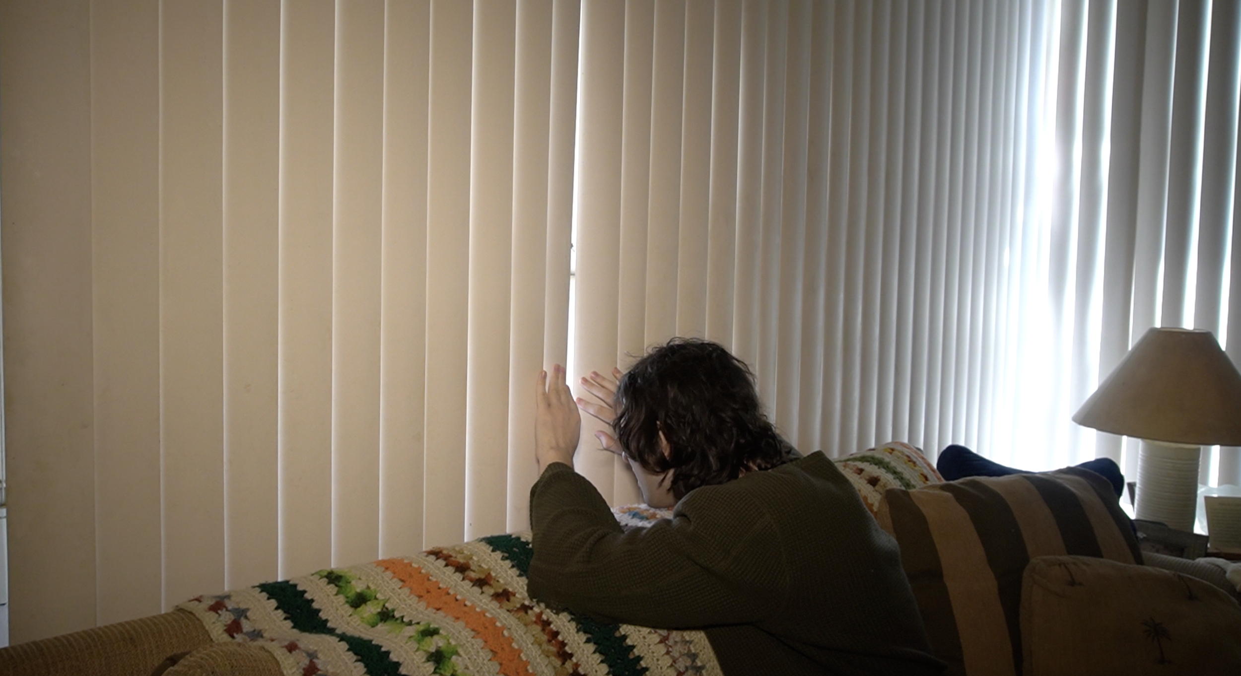 Person with curly dark hair sitting on a sofa, looking out of closed vertical blinds in a living room.