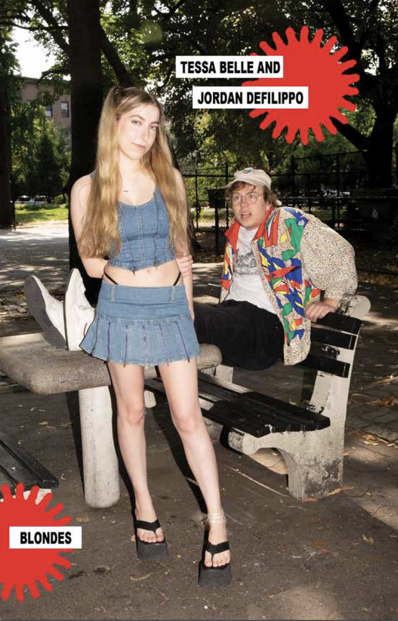 A young woman with long blonde hair wearing a denim crop top, matching denim skirt, and black platform sandals standing in front of a park bench where a young man with curly hair, glasses, colorful jacket, and khaki cap is sitting. The background sho