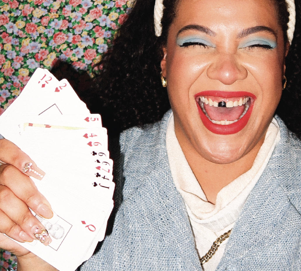 Woman with curly hair, bright makeup, red lipstick, and a headband, smiling widely with a missing front tooth, holding a fan of playing cards, against a floral patterned background.