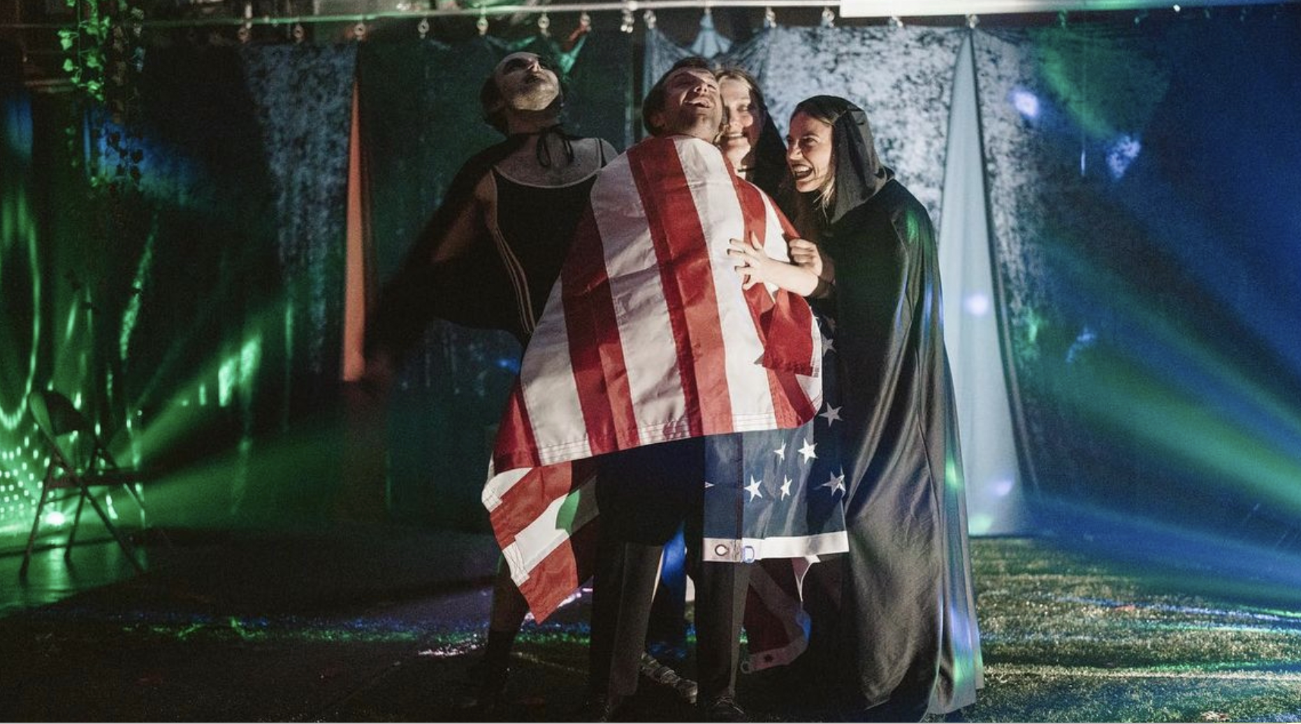 Group of four people dressed in costumes, sharing a joyful moment on a stage with colorful lights and a backdrop.