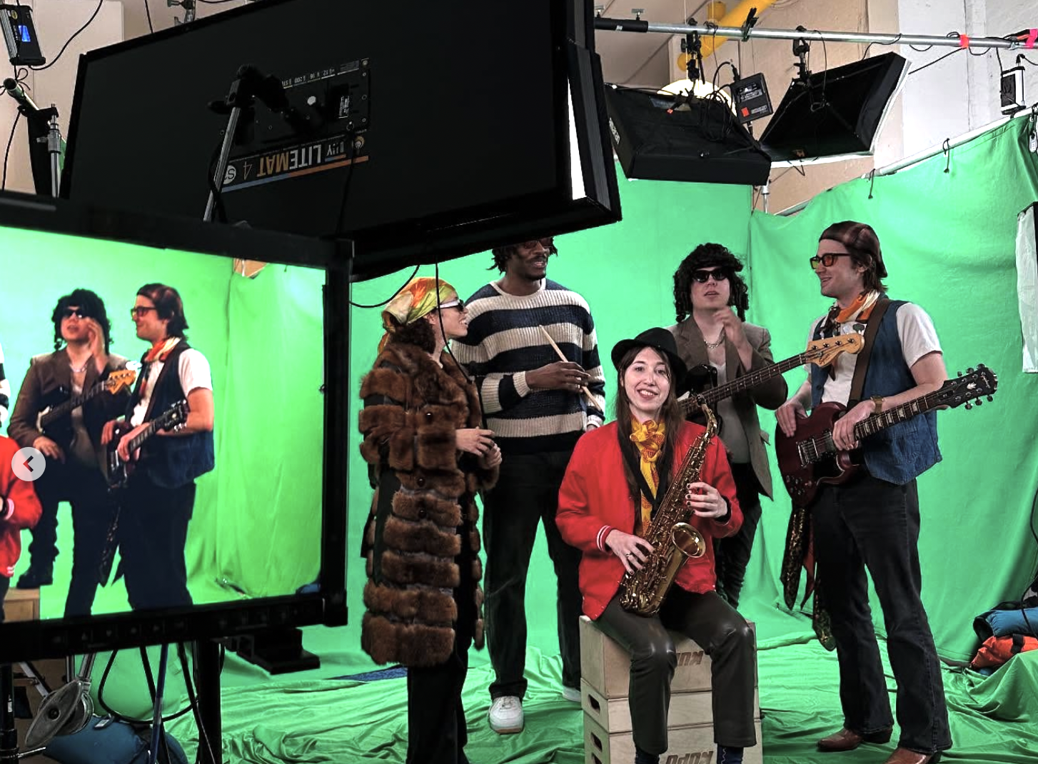 A group of people dressed in retro outfits, including faux fur coats, striped sweaters, and vintage glasses, are in front of a green screen on a film set, with some holding musical instruments like a guitar and a saxophone. A monitor screen shows their reflection.
