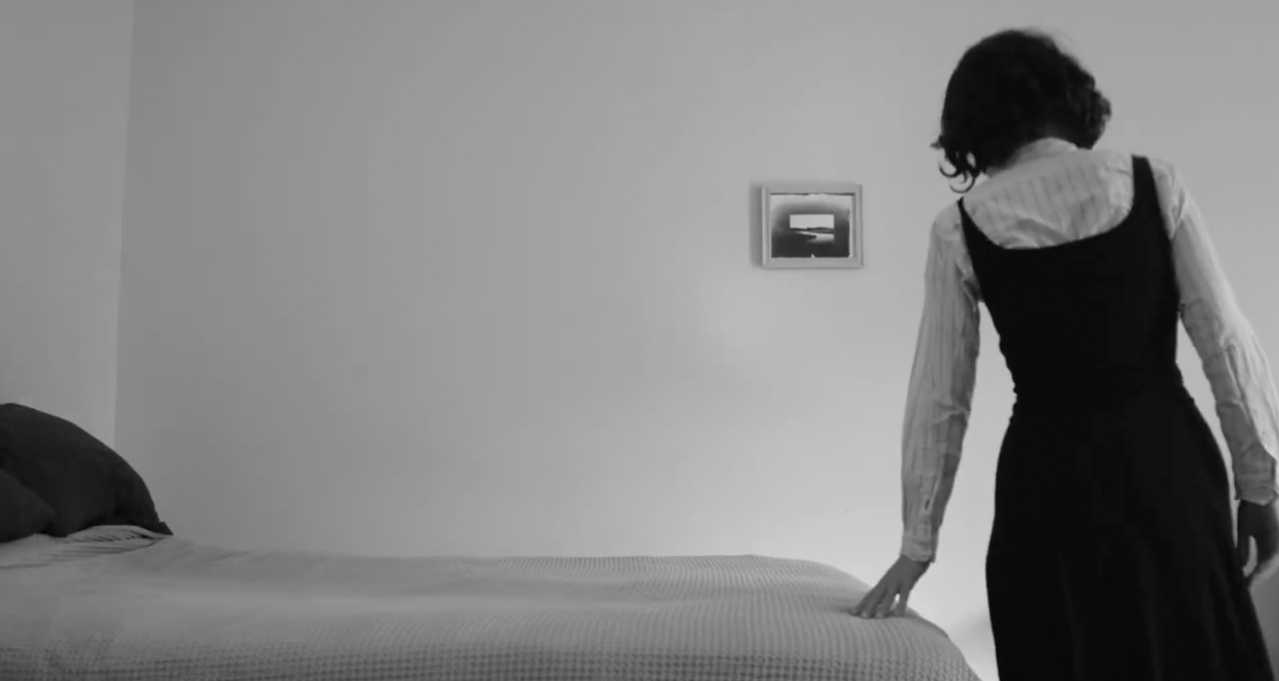 A woman with dark, curly hair, dressed in a striped shirt and black dress, stands beside a bed in a minimalist room, touching the bed frame with her right hand. The room has a plain wall with a small picture hanging above the bed.