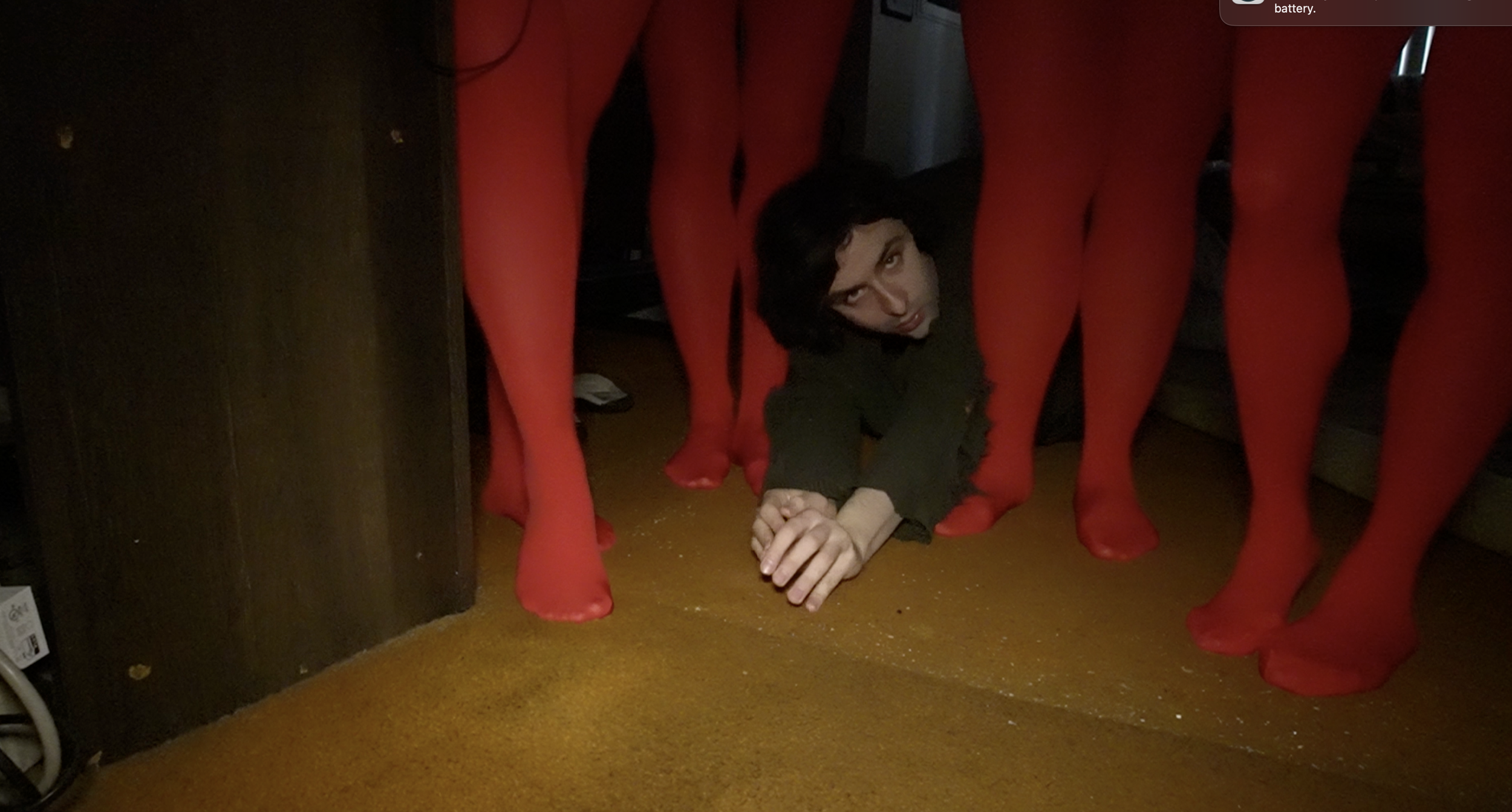 A person with dark hair in a black shirt is underneath a group of women wearing red tights, with only their legs visible. The person is lying on the floor, looking up through the legs.