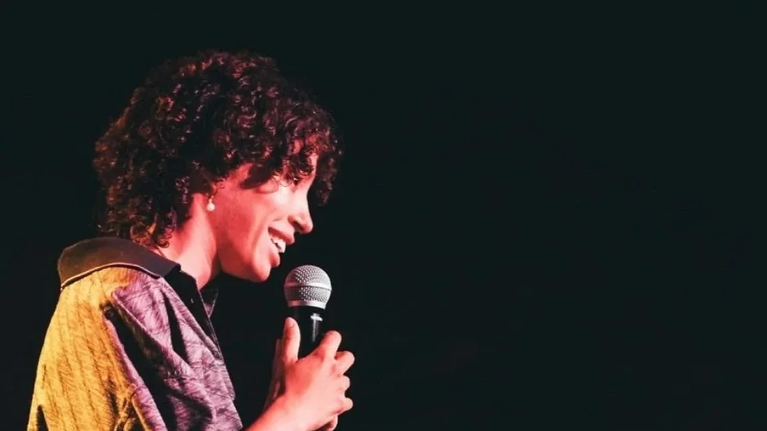 A woman with short curly hair smiling and speaking into a microphone on a dark stage.