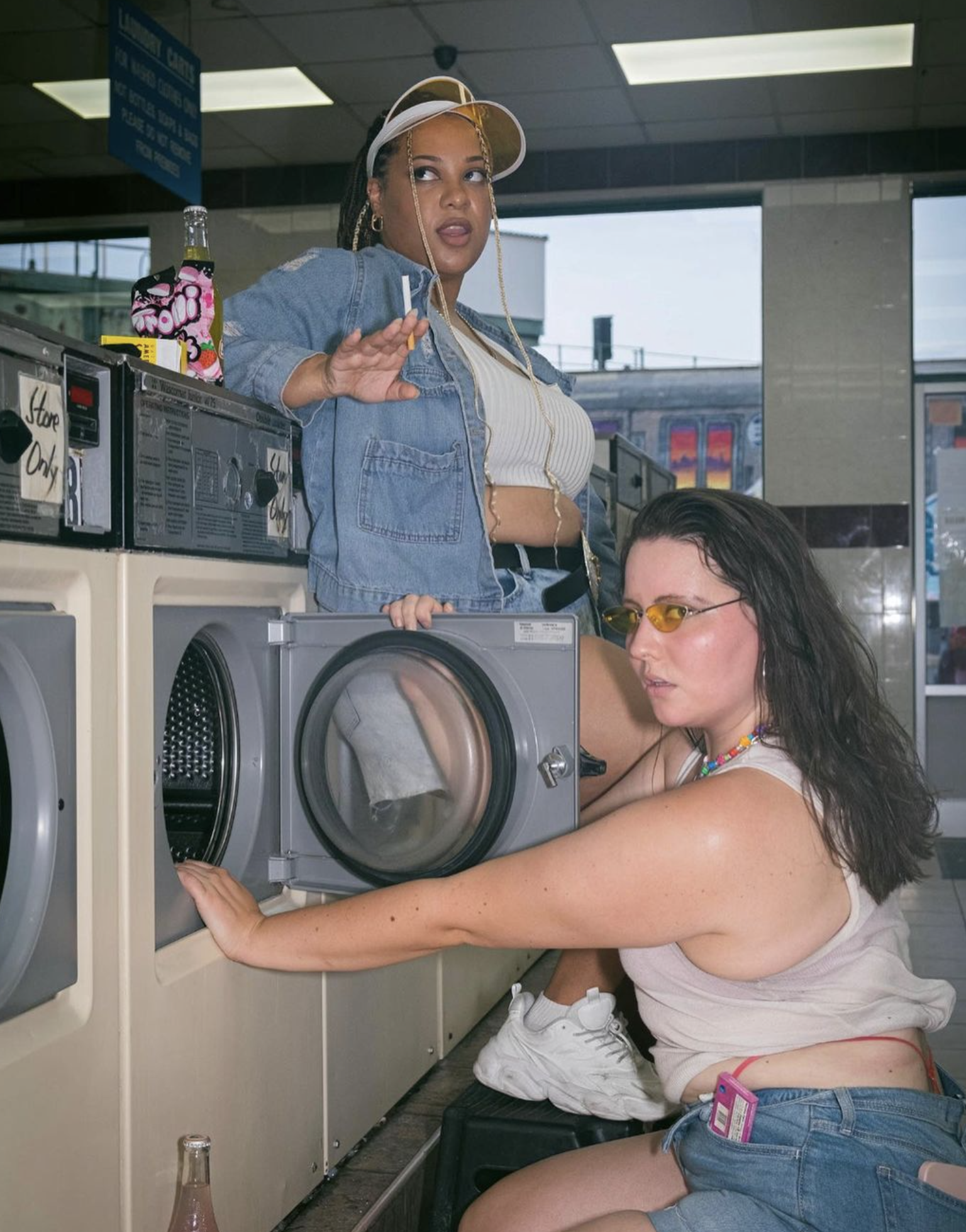 Two women are in a laundromat, one is standing and the other is crouched next to a washing machine. Both women are wearing sunglasses and casual clothing, with the crouching woman holding the door of the washing machine open.