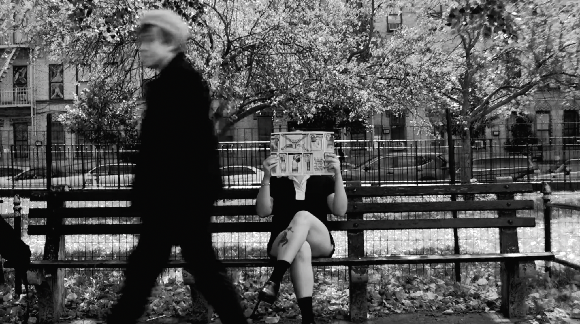 Black and white photo of a park with blooming trees, featuring a person sitting on a park bench reading a comic book, and a blurred person walking past in the foreground.