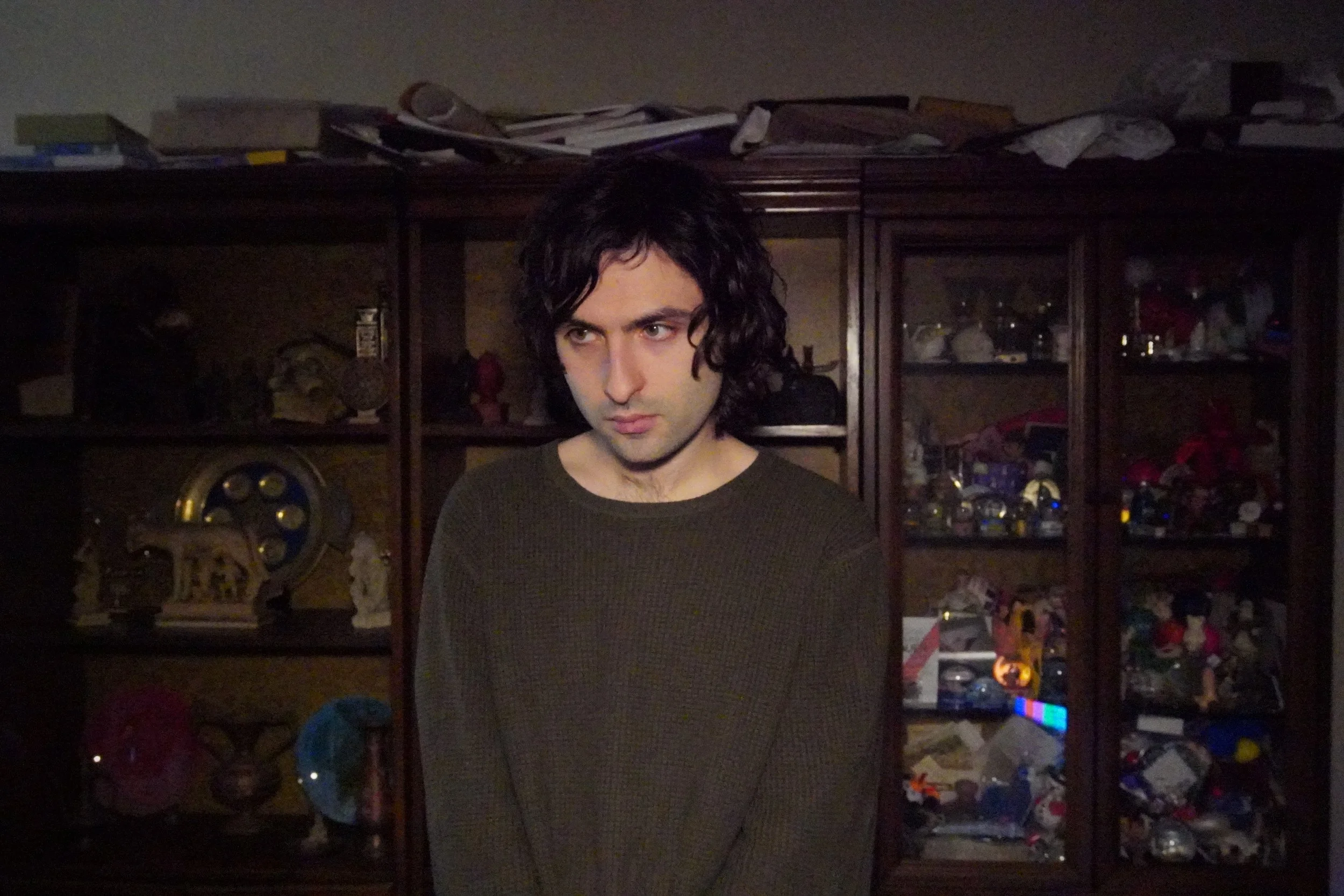 A man with dark, curly hair and wearing a brown sweater stands in front of a dark wooden display case filled with souvenirs and collectibles, in a dimly lit room.