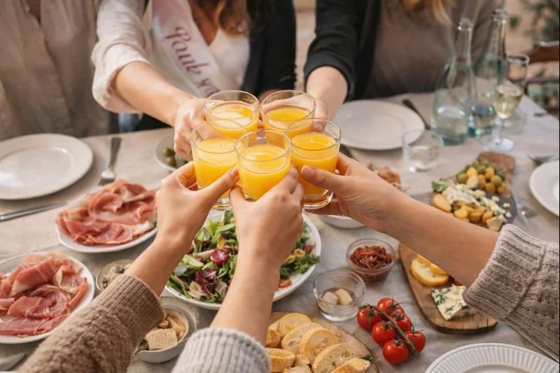Brunch tra amiche per un addio al nubilato, brindisi con succhi e piatti condivisi in un momento conviviale.