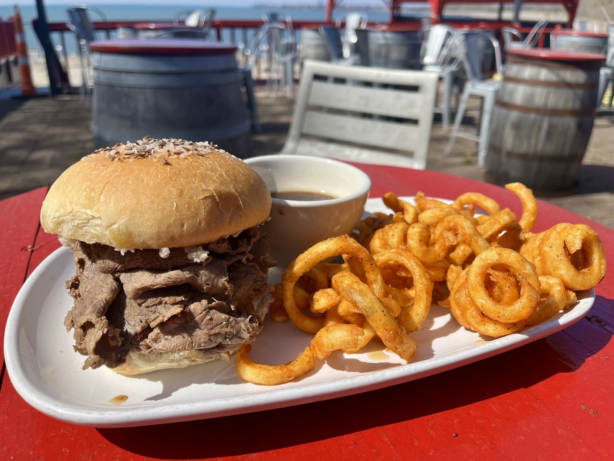 A plate with a sandwich containing shredded beef, curly fries, a small cup of dipping sauce, on a red table outdoors with barrels and chairs in the background.