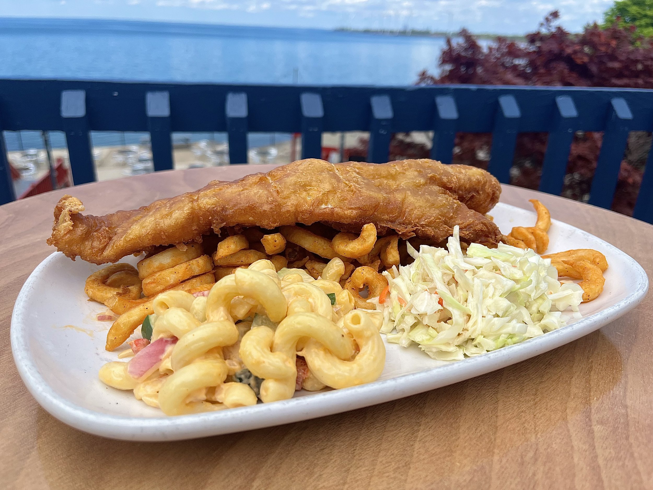 Fried fish fillet with curly fries, macaroni salad, and coleslaw on a white plate outdoors with a body of water and trees in the background.