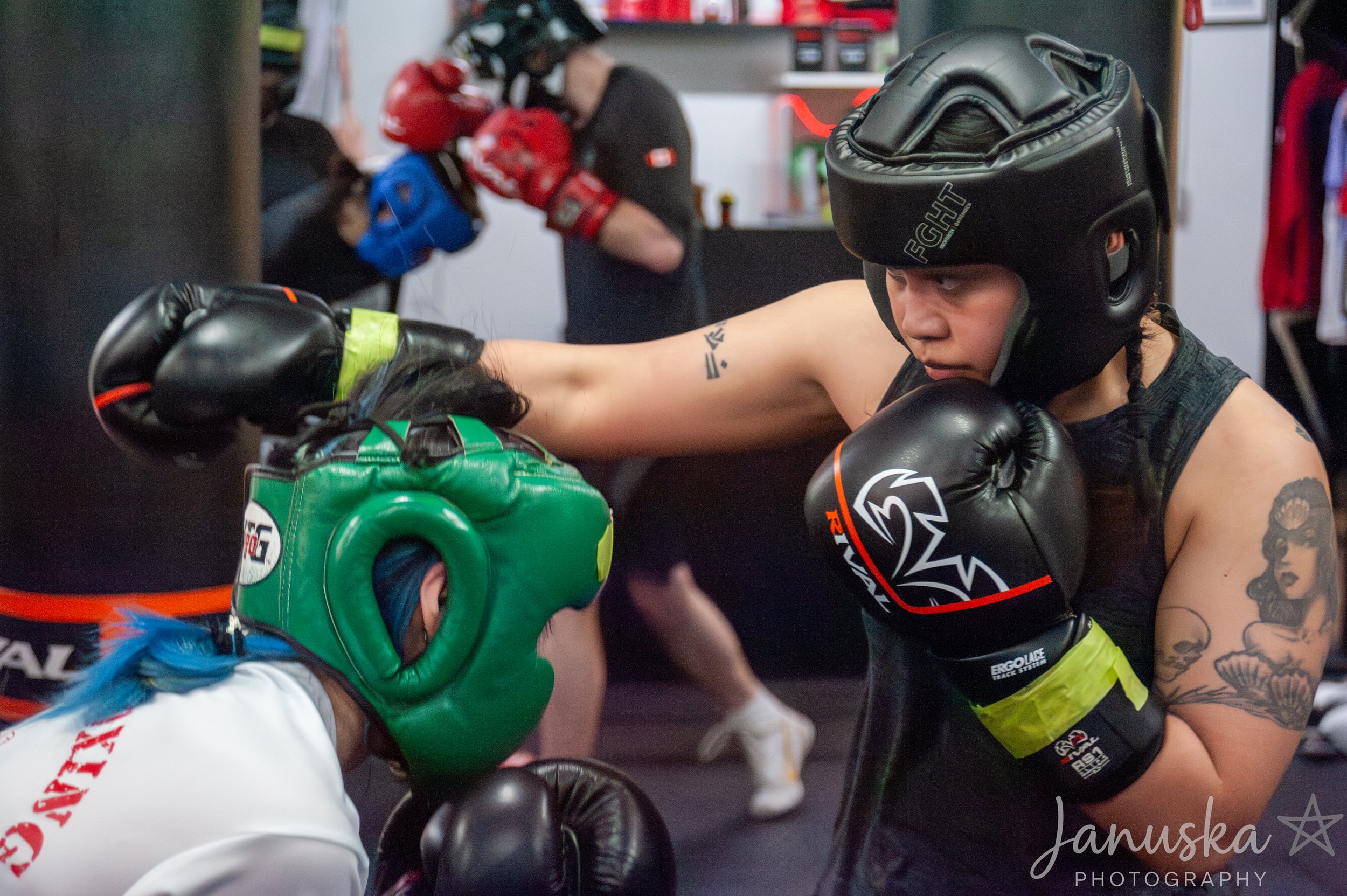 Two women wearing boxing gear, including helmets and gloves, are training in a boxing gym. One woman is in focus on the right, with tattoos on her arm, preparing to punch. The other woman, wearing a green headgear, is in front of her, holding boxing gloves. In the background, a woman with a red helmet is practicing with a punching bag.