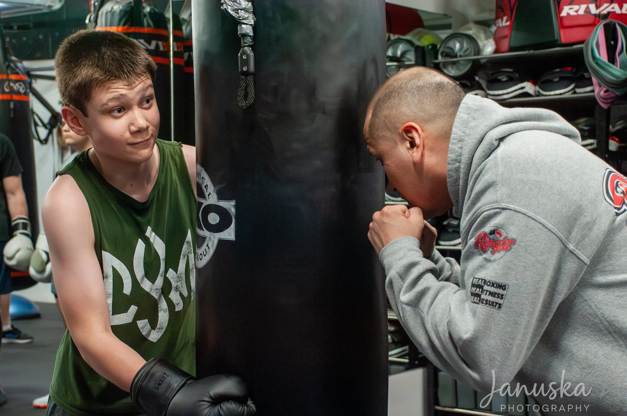 A boxing gym scene with a young male boxer wearing black boxing gloves and a green sleeveless shirt, standing next to a black punching bag, and an adult male trainer wearing a grey hoodie and boxing shorts, leaning in with his hands clasped. The background shows shelves with boxing equipment and other gym items.