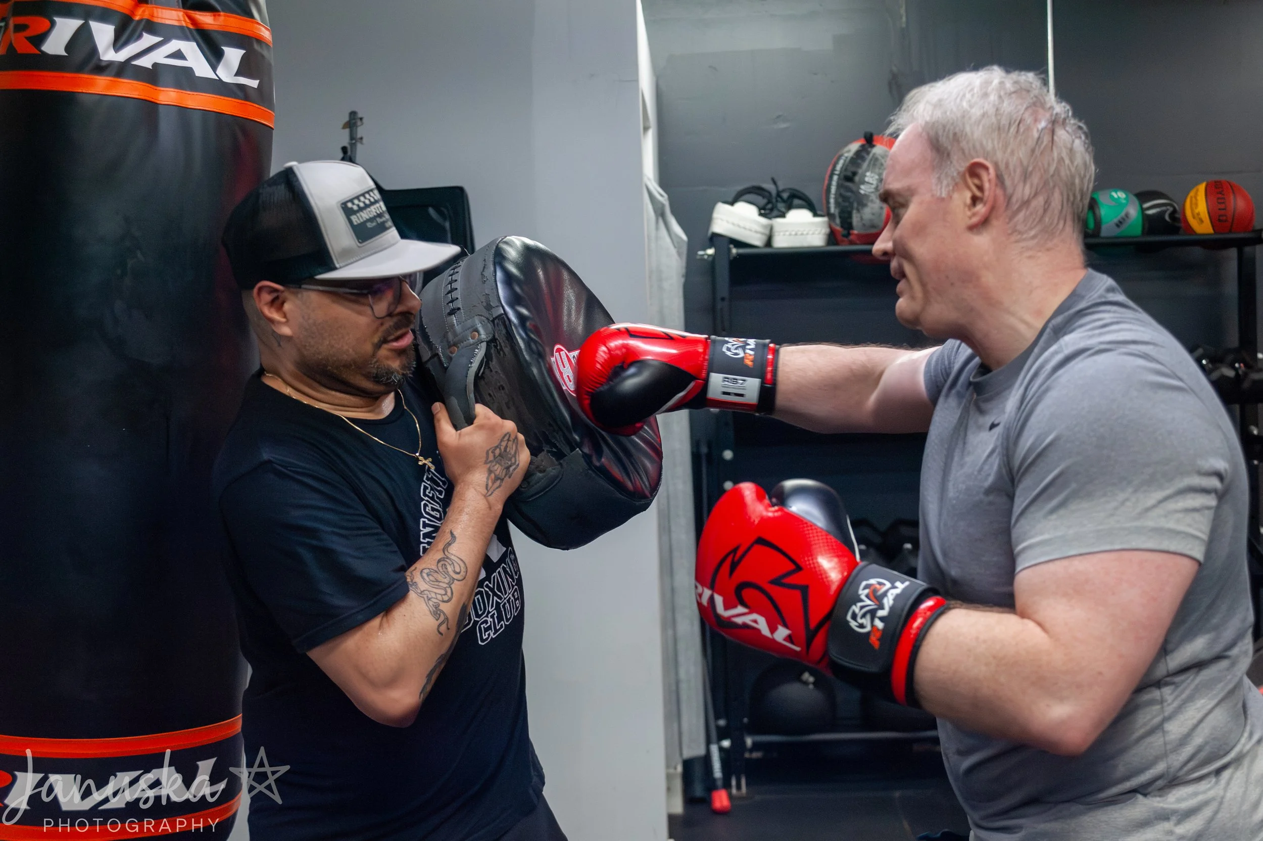 Two men are in a boxing gym. One man, wearing boxing gloves and a gray t-shirt, is punching a black boxing pad held by a woman. The woman, wearing glasses, a black t-shirt, a cap, and a gold necklace, is holding the pad and assisting the boxer.