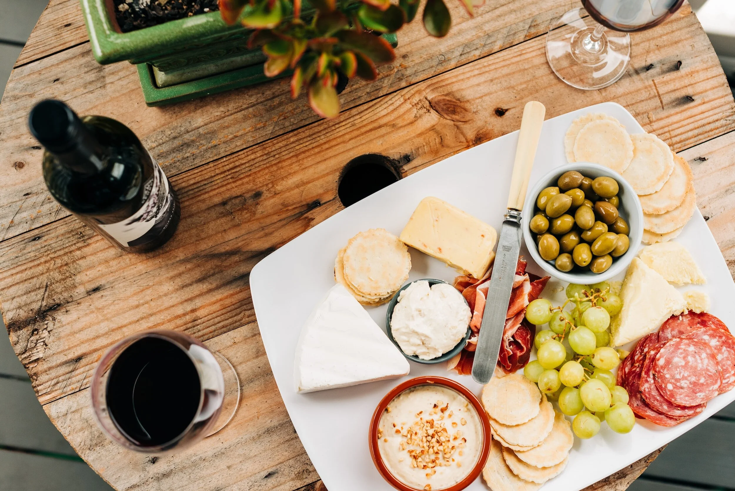 A cheese and charcuterie board with grapes, olives, crackers, and wine glasses on a wooden table.