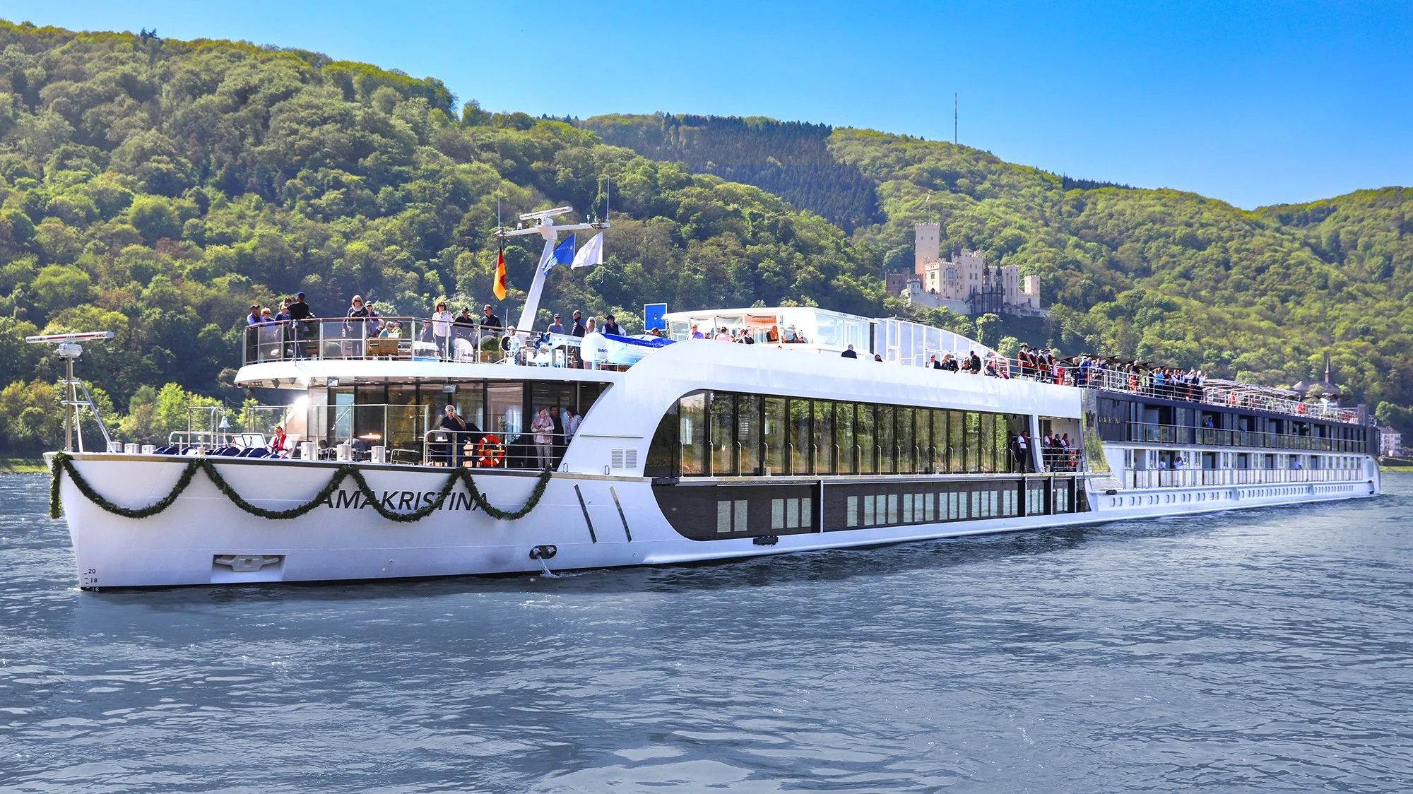 A large, modern white cruise ship with multiple decks sailing on a river, decorated with greenery, with many people on the decks enjoying the scenery. A castle is visible on a hill in the background.