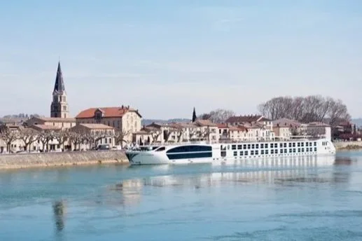 A large, modern white cruise ship with multiple decks sailing on a river, decorated with greenery, with many people on the decks enjoying the scenery. A castle is visible on a hill in the background.