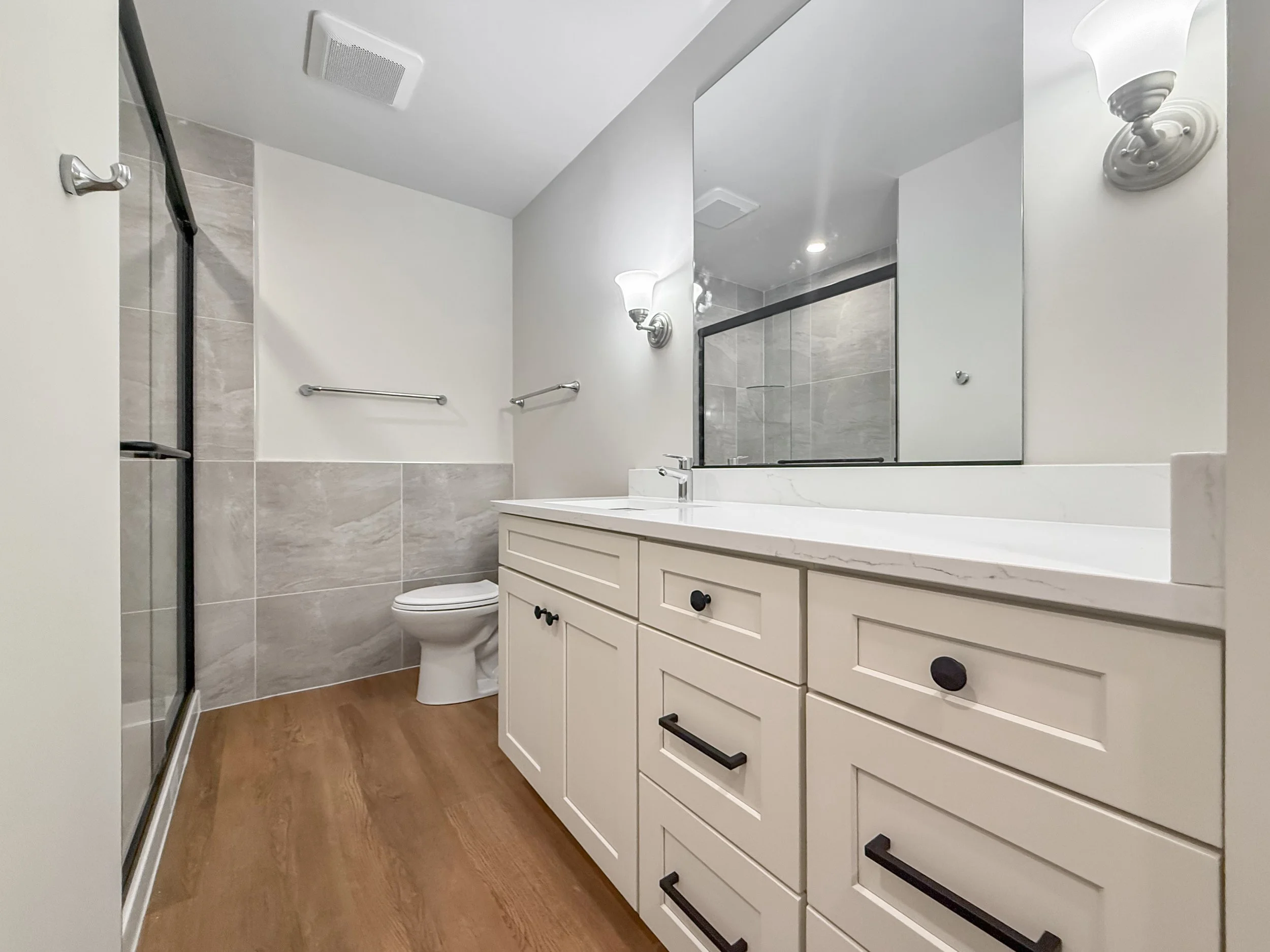 Modern bathroom featuring a beige vanity with black hardware, a large mirror, wall-mounted light fixtures, a toilet, a walk-in shower with glass door, and wood-style flooring.