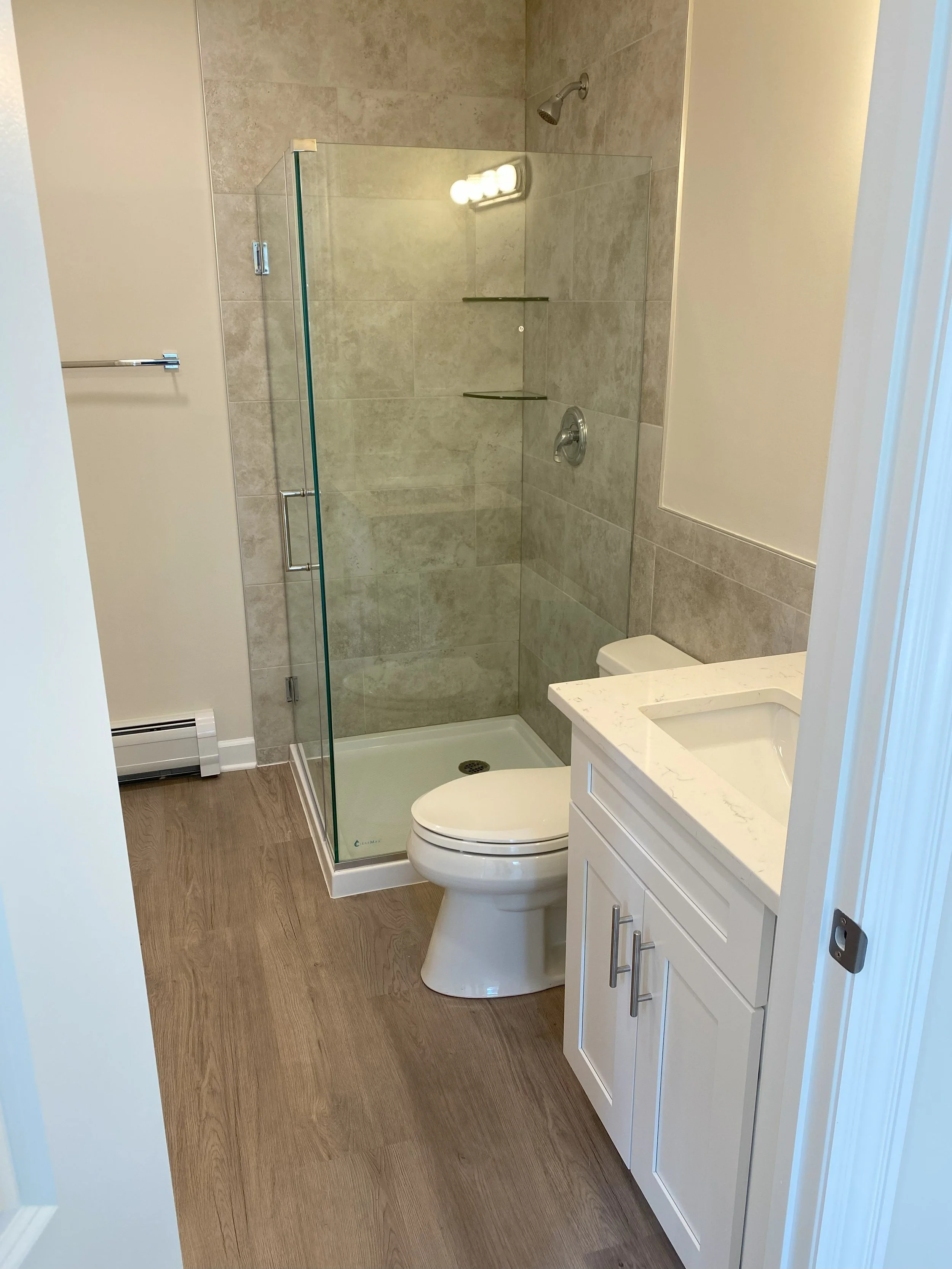 Small bathroom with a wooden floor, white vanity with a marble countertop, toilet, and glass-enclosed shower with beige tiles and built-in shelves.