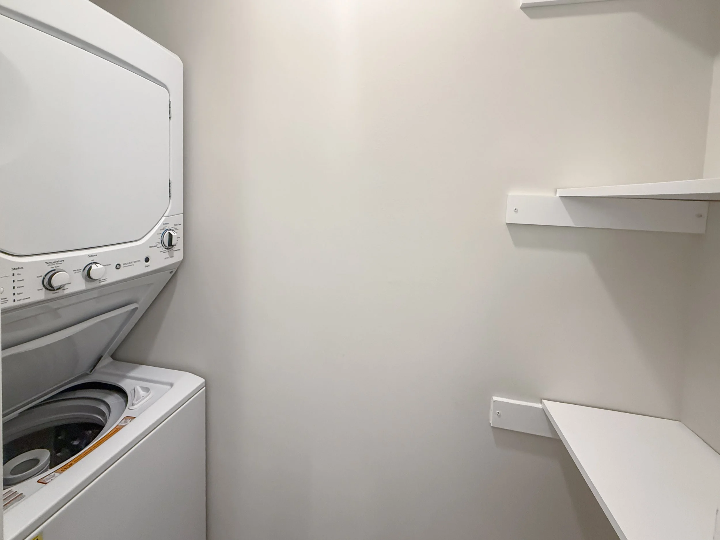 A white stacked washer and dryer unit in a small laundry room with empty white shelves on the right side.