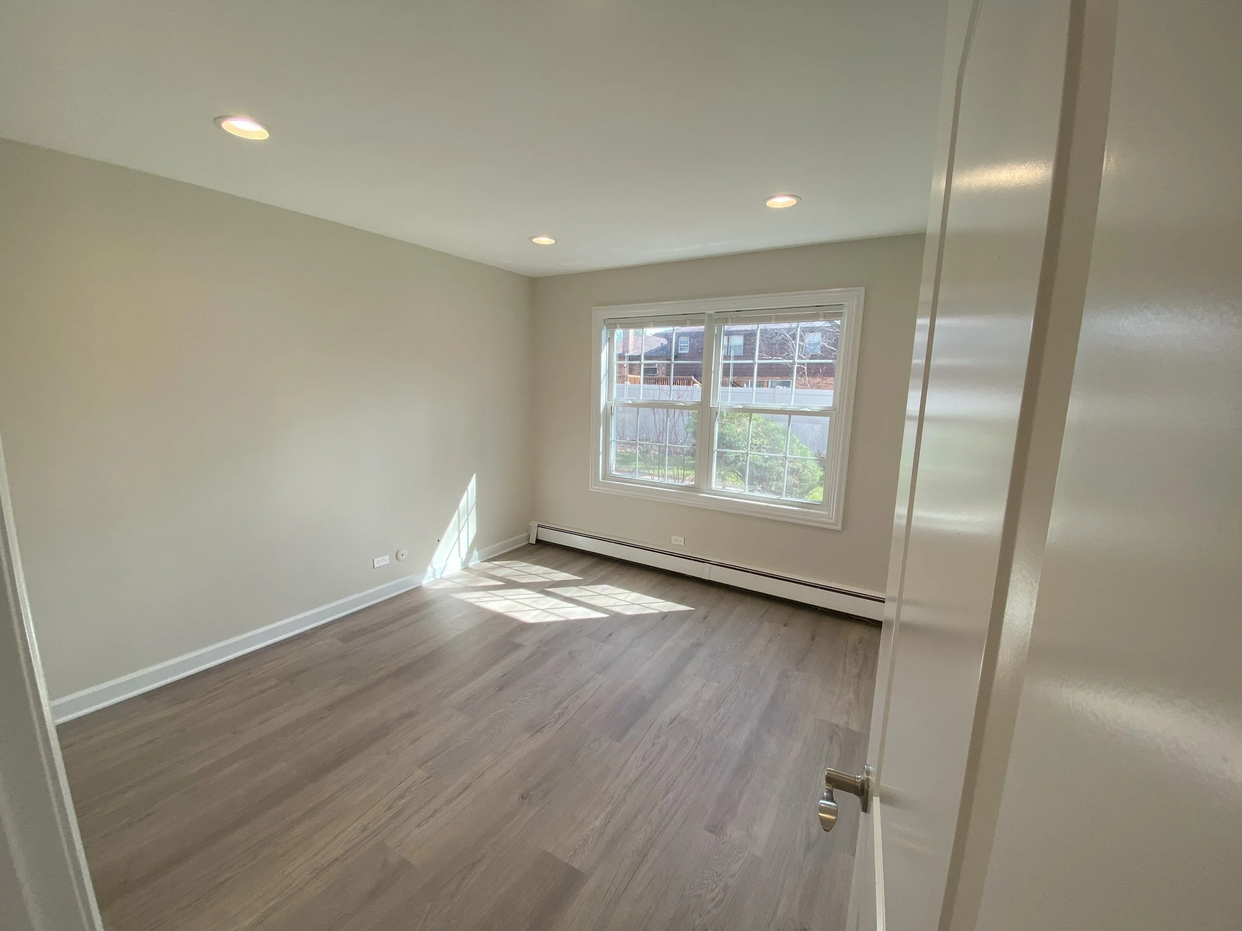 Empty room with beige walls, hardwood floor, large window letting in natural light, and recessed ceiling lights.