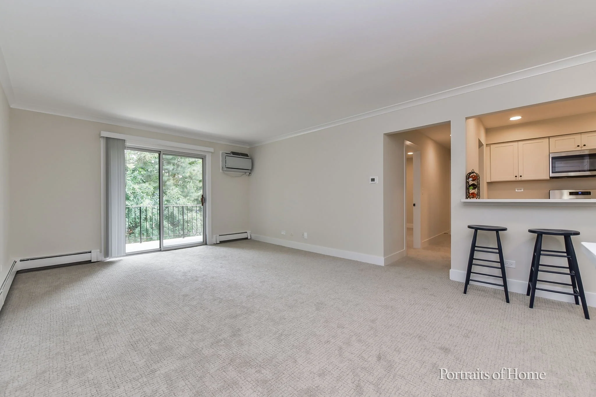 Empty living room with beige carpet, sliding glass door leads to balcony, window air conditioning unit, open kitchen area with breakfast bar, two black barstools, white cabinetry, and stainless steel appliances.