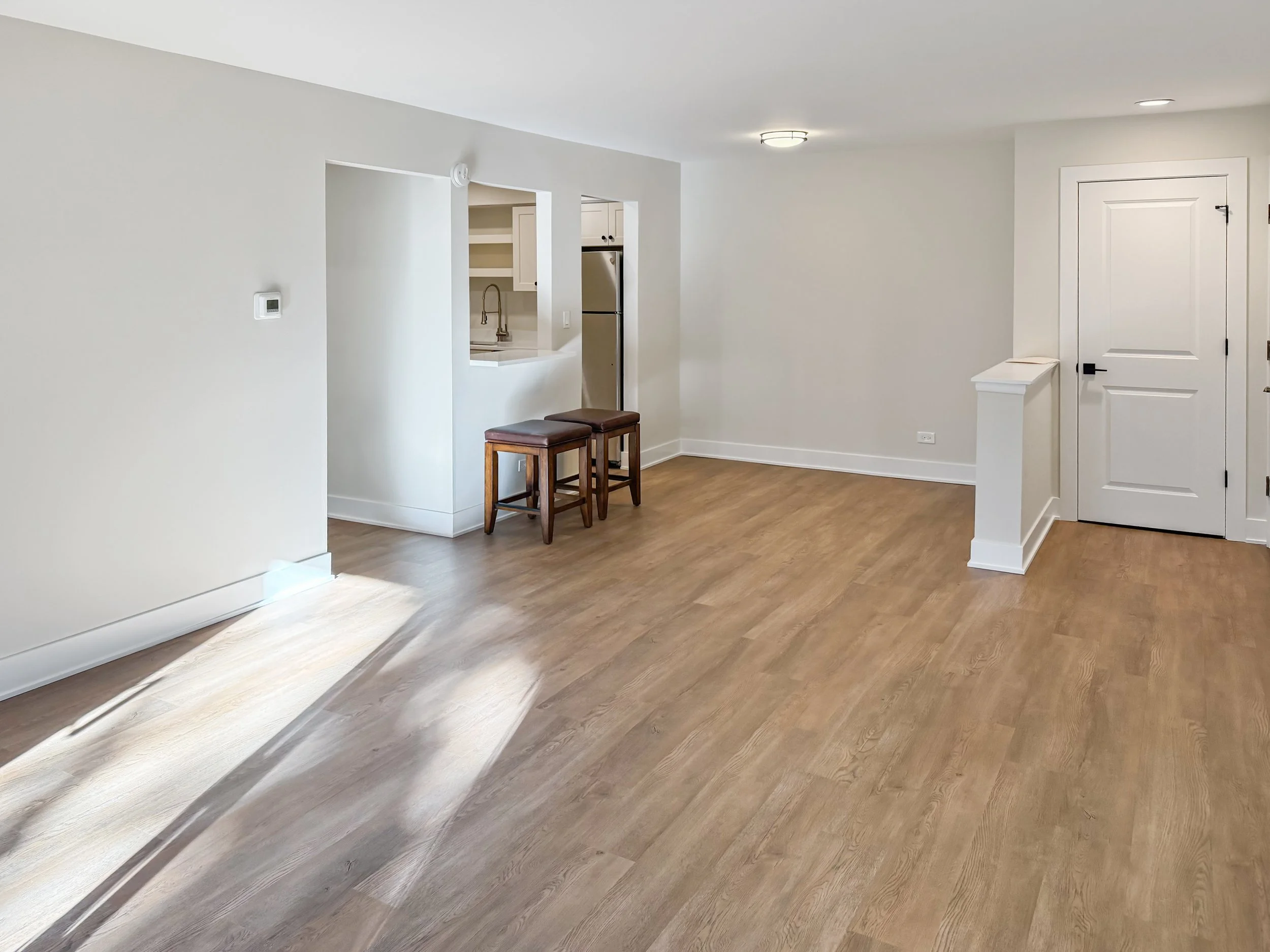 Empty living room with light wood flooring, white walls, a small kitchen area with a pass-through window, and two bar stools. There is a door on the right side, and ceiling lights are visible.