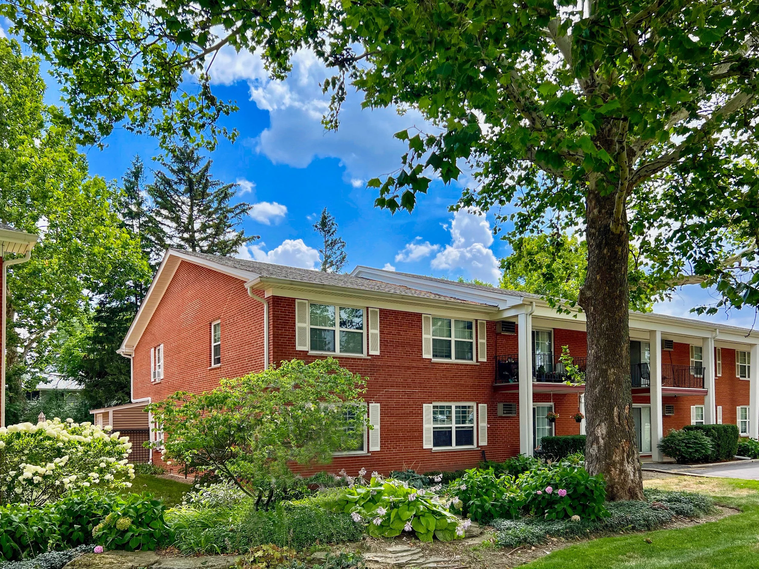 A red brick residential apartment complex with white trim, surrounded by green trees and landscaping, under a blue sky with clouds.