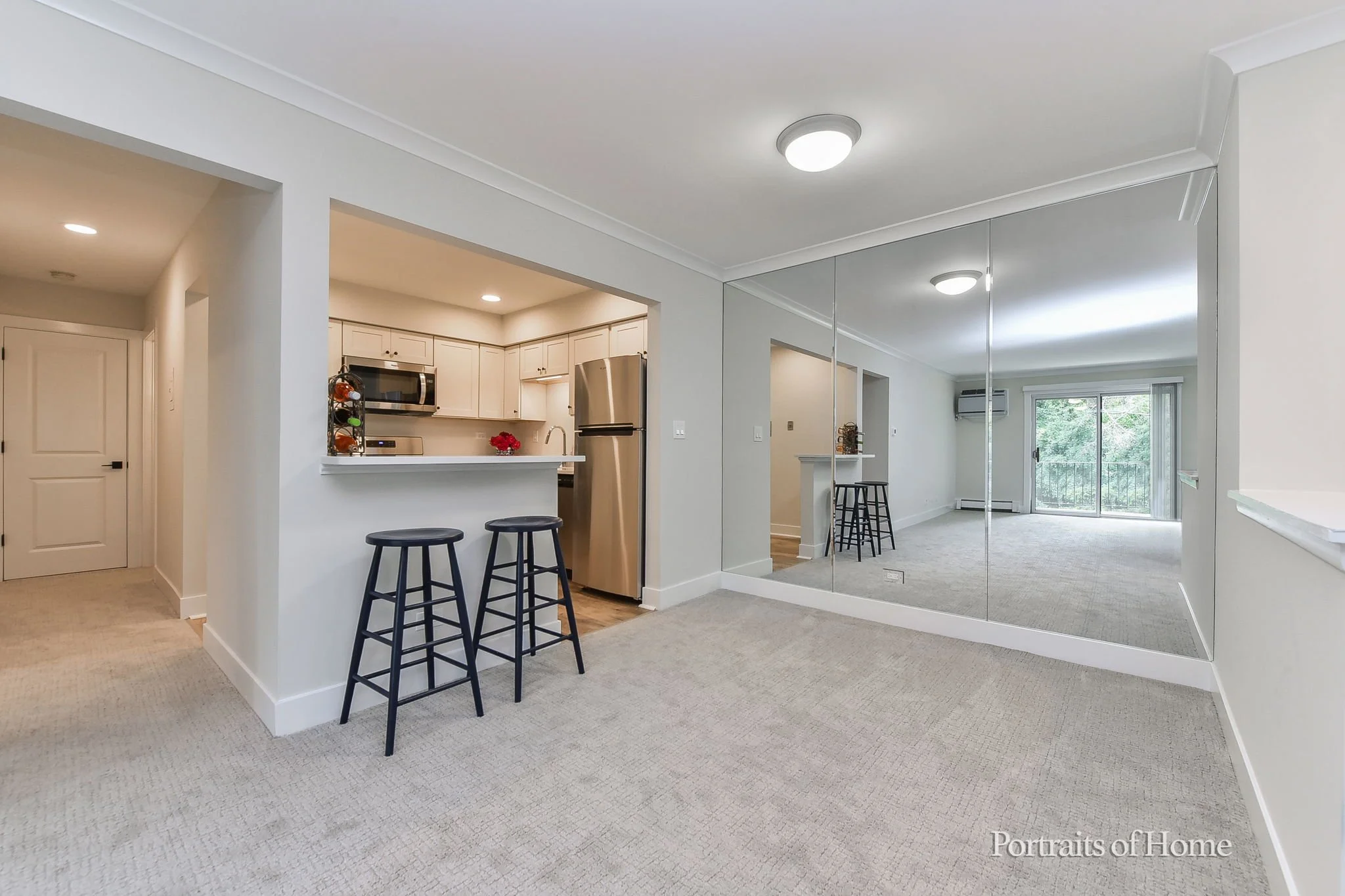 Living space with a view of the kitchen, featuring a breakfast bar with two black stools, a large mirror on the wall, and a sliding glass door leading to a balcony with trees outside.