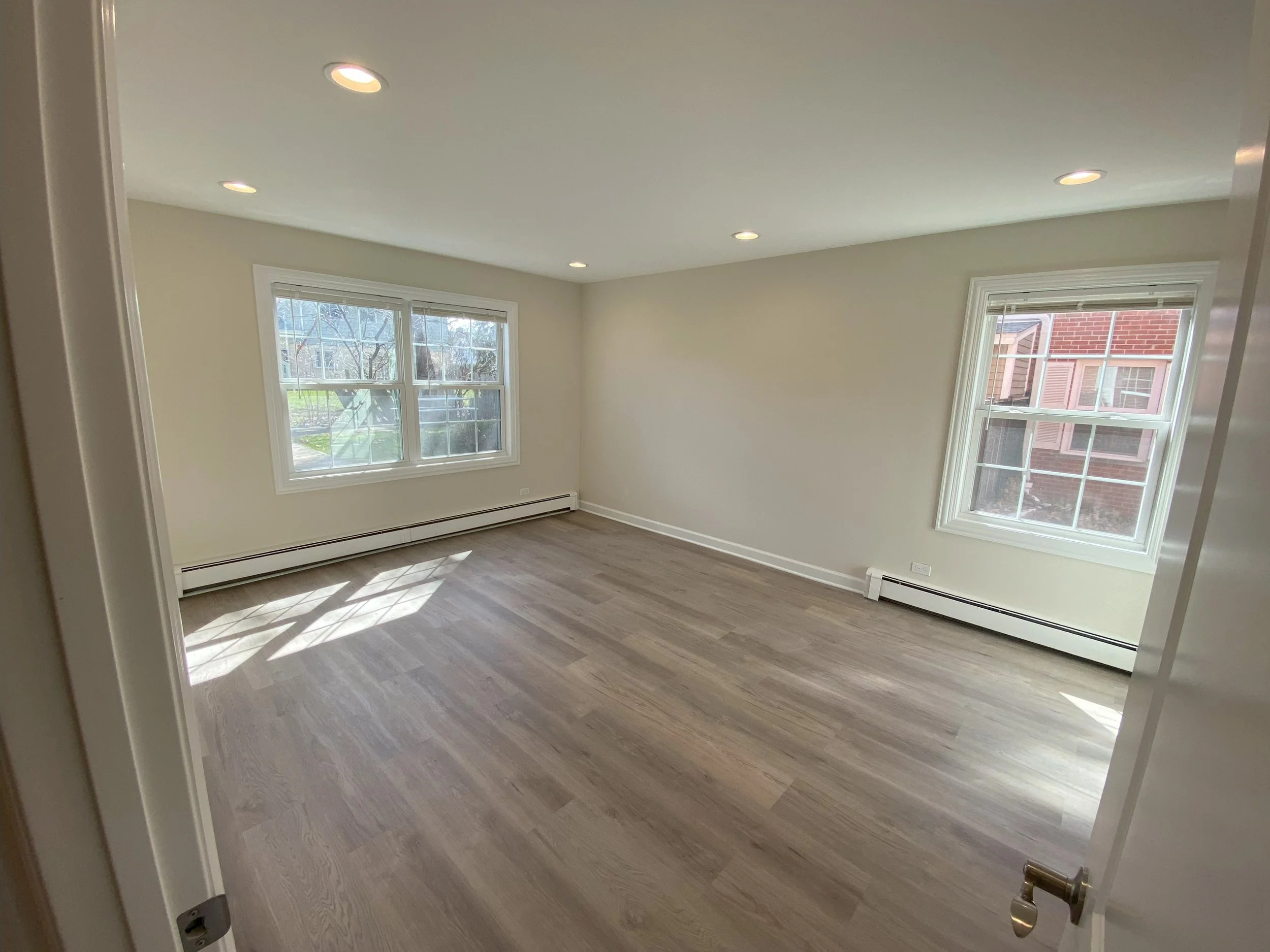 Empty room with hardwood floors, white walls, three windows letting in natural light, and recessed ceiling lights.