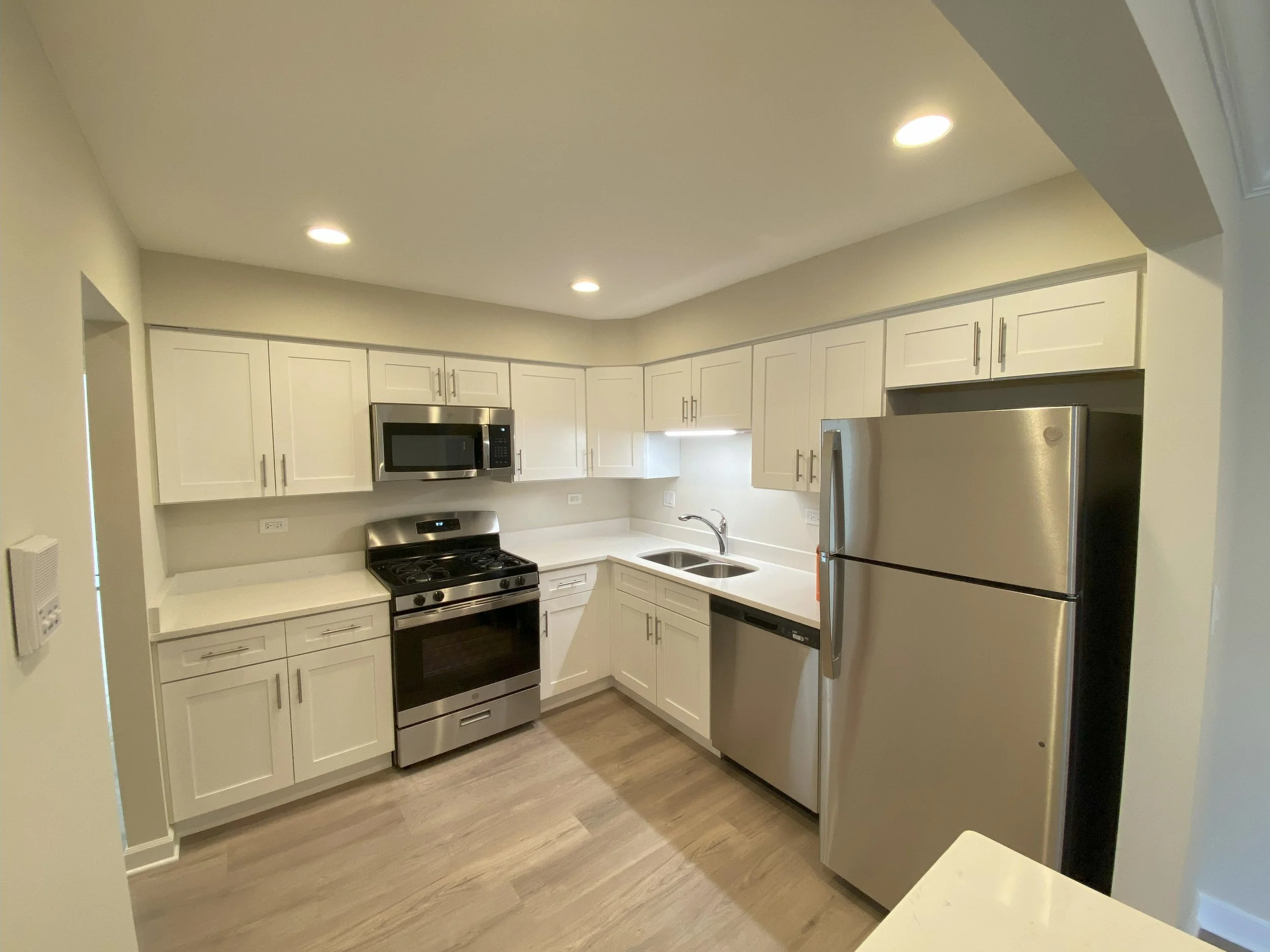 Modern kitchen with white cabinets, stainless steel fridge, microwave, and stove, light wood flooring, and a small sink under a window.