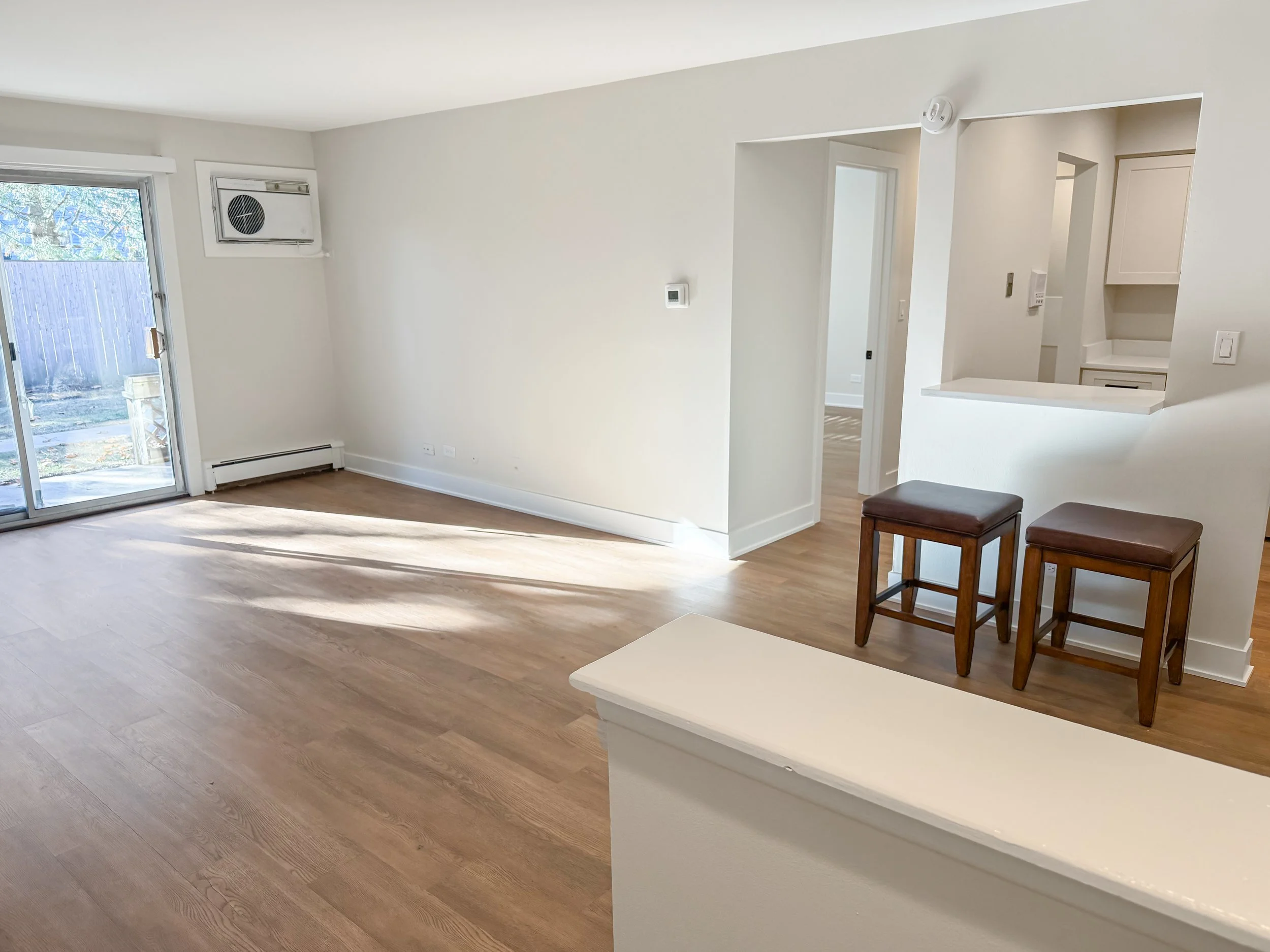 Empty living room with wooden flooring, a sliding glass door leading outside, and a small opening in the wall revealing a kitchen. Two brown barstools are placed against the wall.